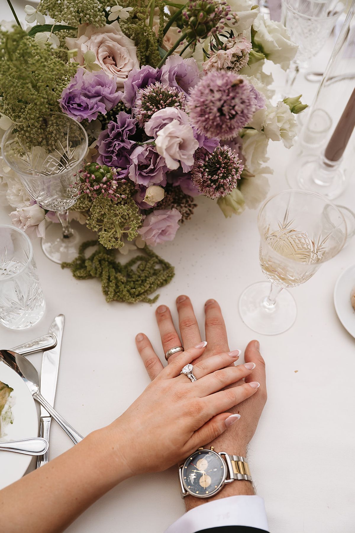Two hands rest together on a white tablecloth, showing rings and a wristwatch, beside lilac flowers and wine glasses.