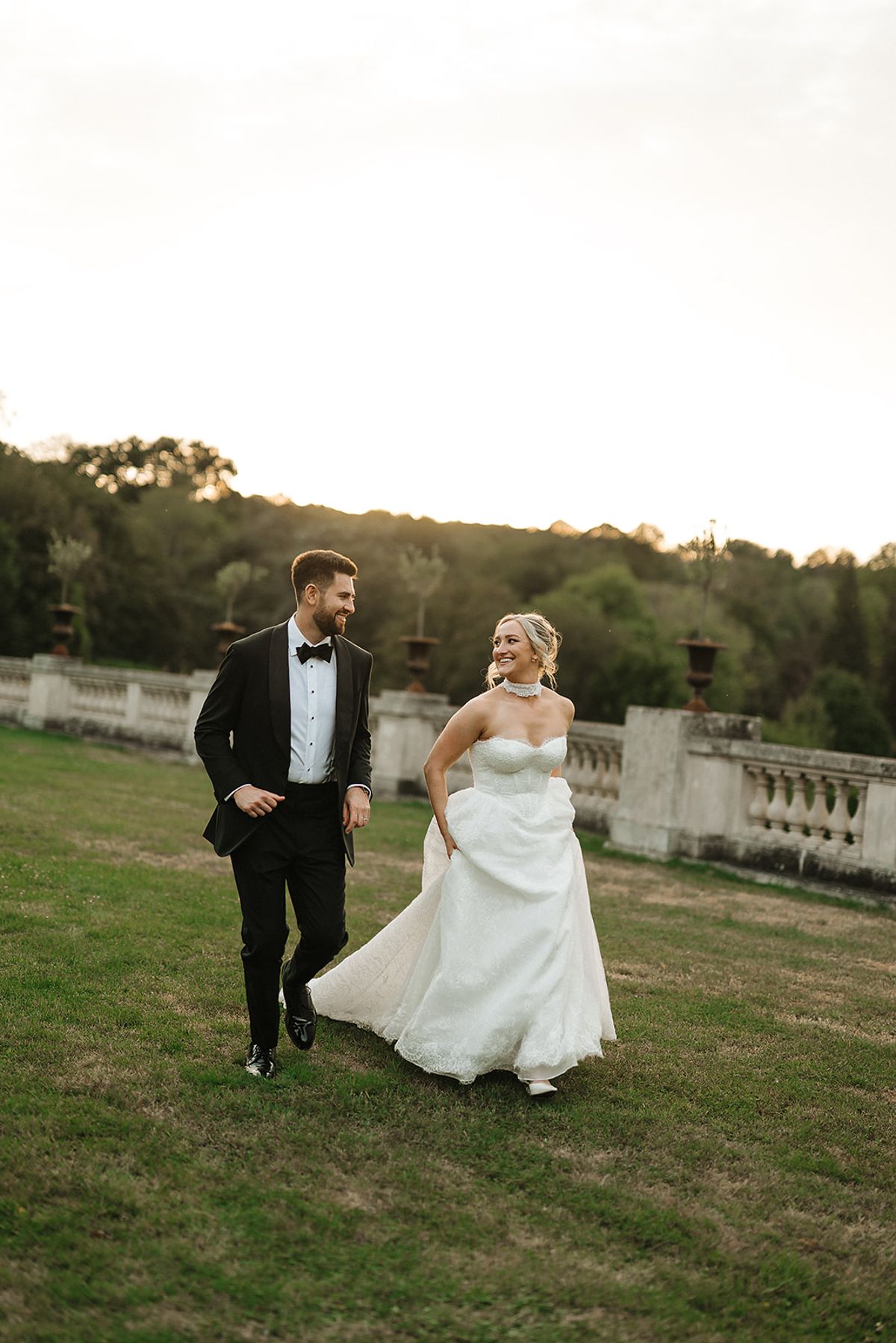 The groom in a dark suit and the bride holding up her white gown walk across a lawn towards the camera, smiling.