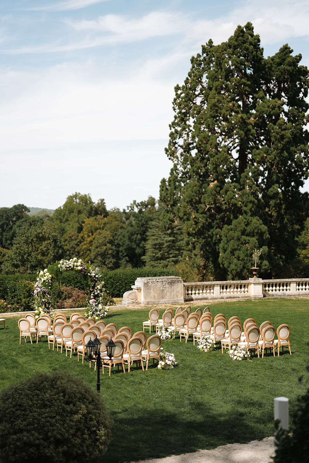 Rows of wooden chairs and a floral arch stand on a manicured lawn beside a tall tree and a stone balustrade.