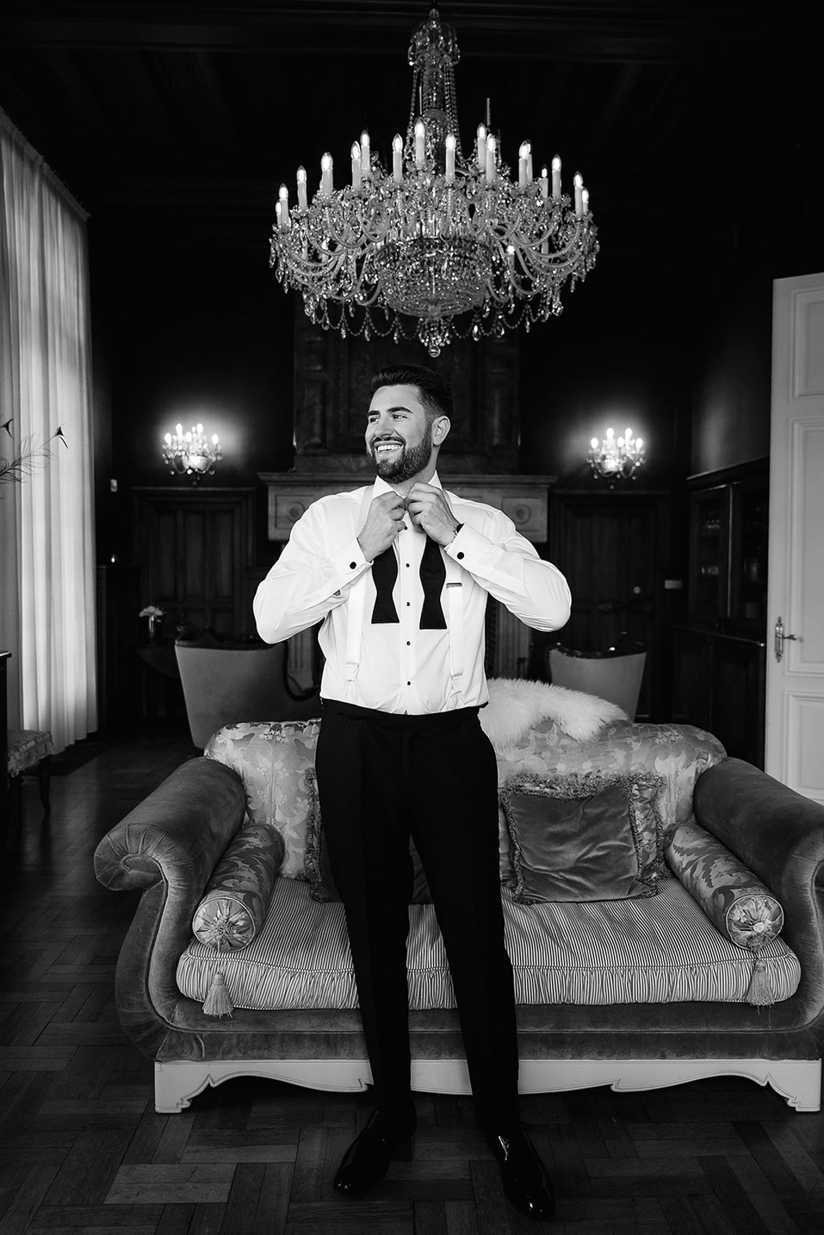In black and white, the groom in a white shirt and dark trousers adjusts his bow tie in front of a sofa beneath a chandelier.