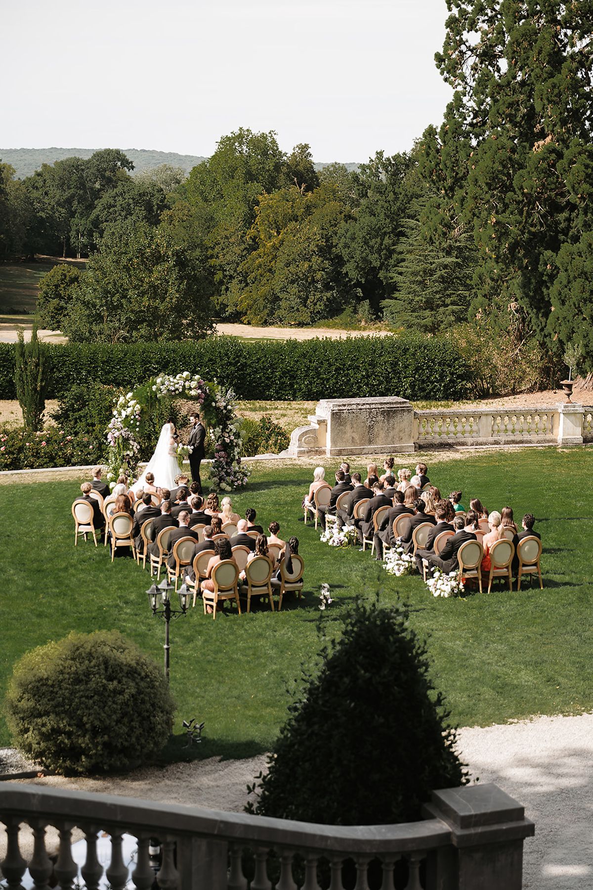 The bride and groom stand before a floral arch while seated guests watch, on a lawn in a large green park.