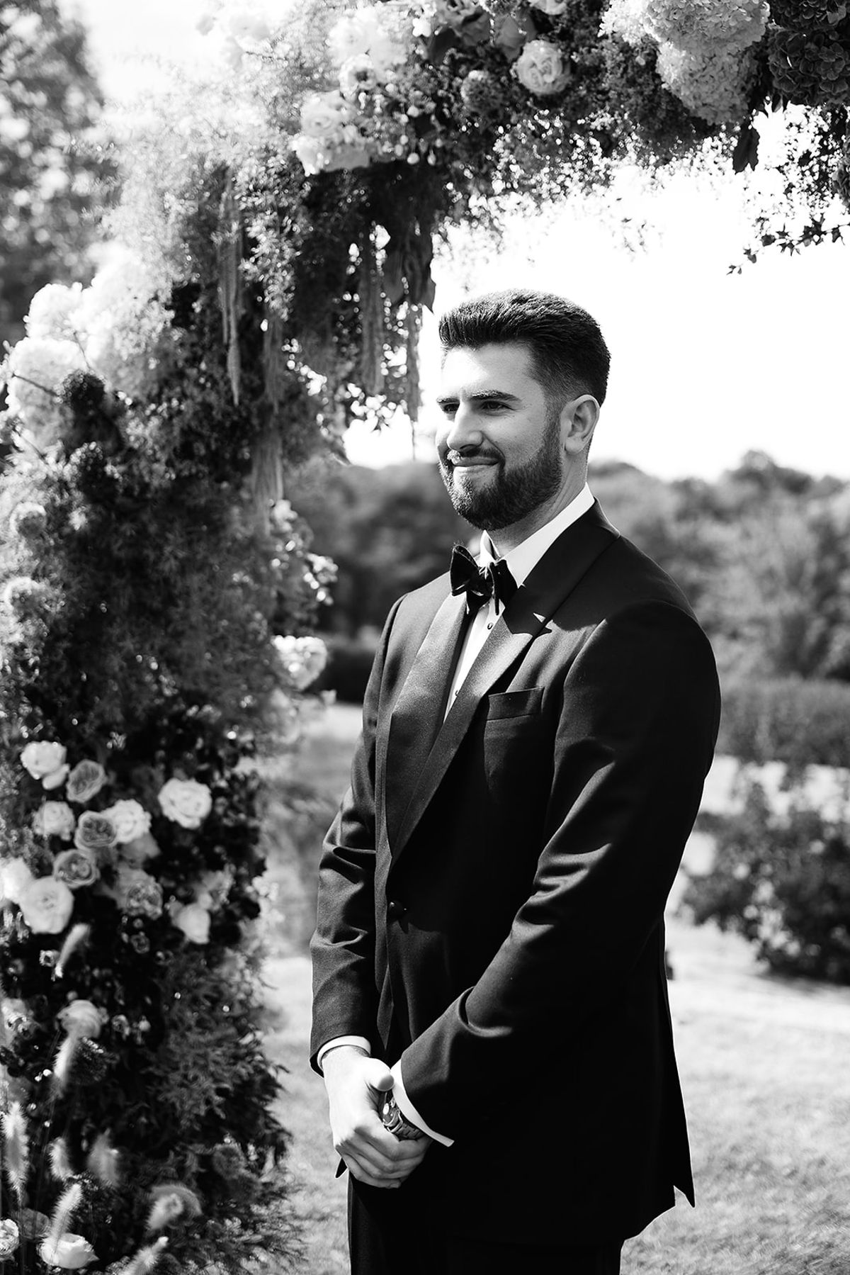 The groom, bearded and in a dark tuxedo and bow tie, smiles beside a flower-covered arch in this black-and-white photo.