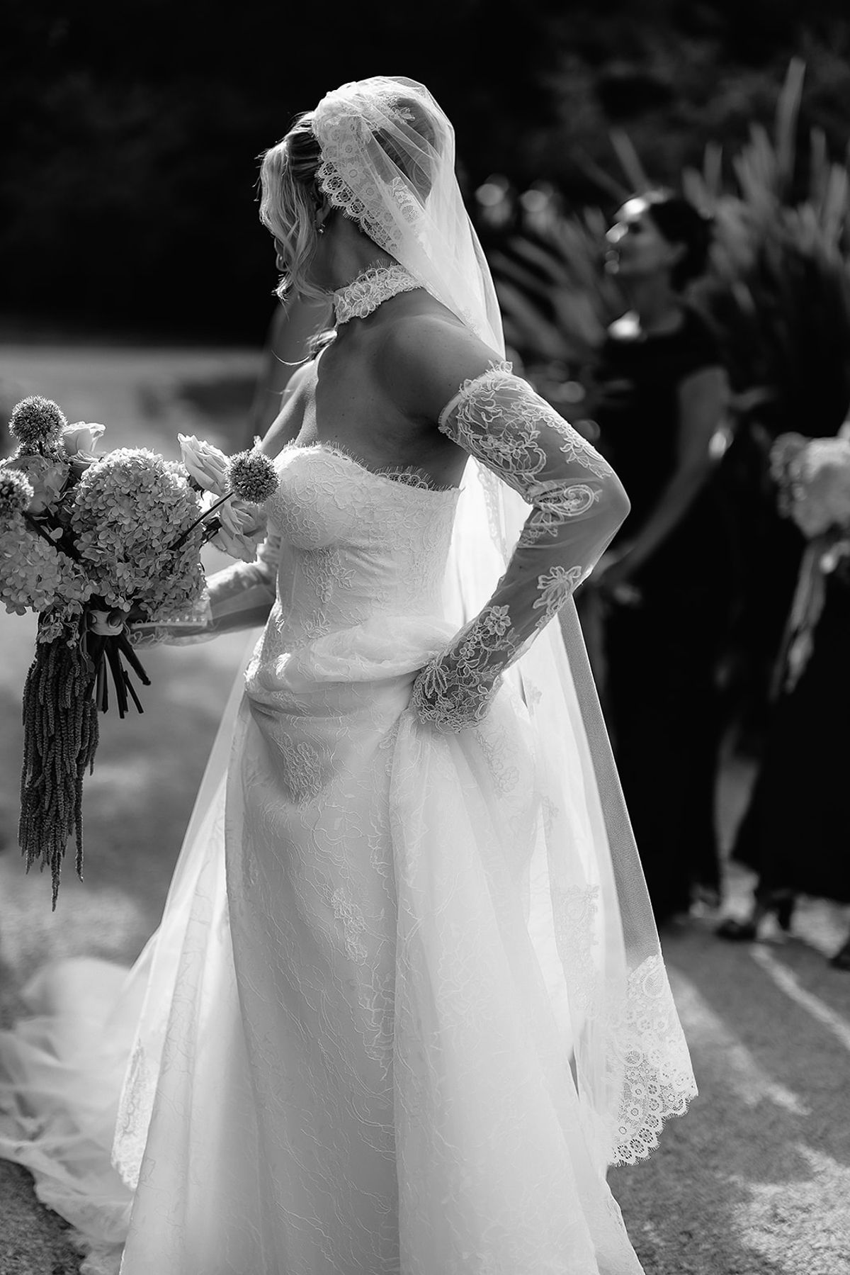 The bride, in a lace gown, long lace gloves and veil, holds a large bouquet while looking away; guests stand behind her.