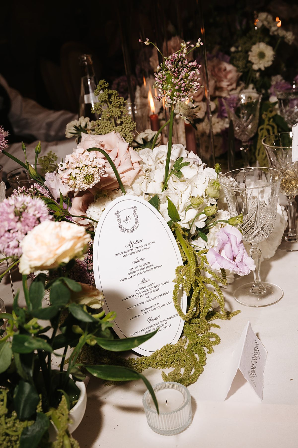 An oval menu card stands among pale pink and white flowers, trailing greenery and crystal glasses on a white-clothed table.