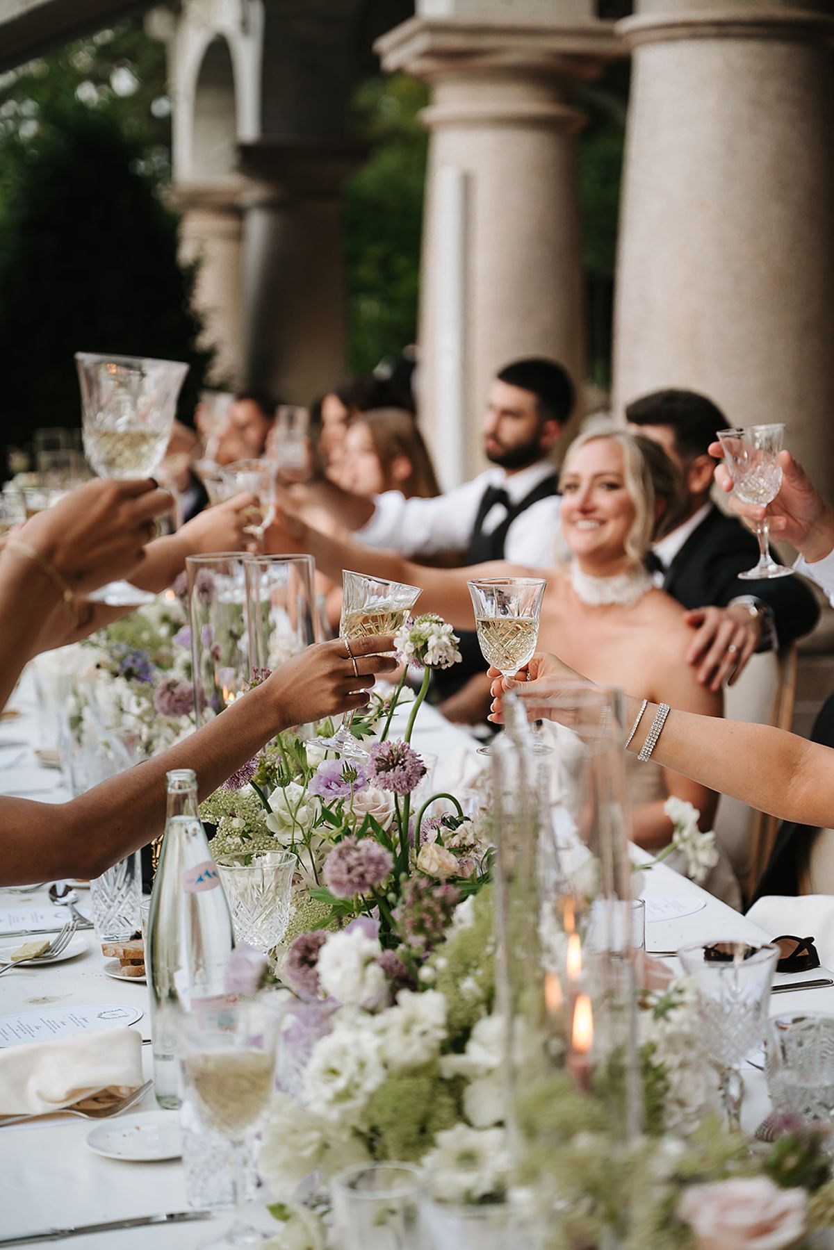 Guests at a long table clink glasses beside tall stemmed flowers and candles, with the bride smiling among them.