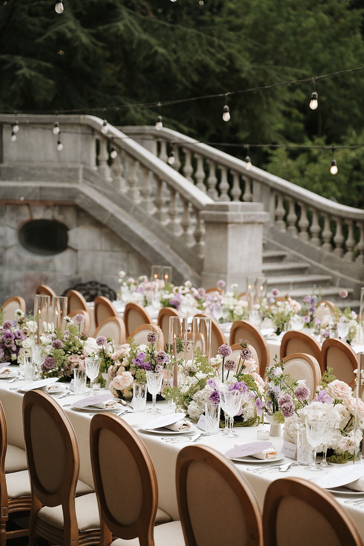 A long table with cream linen, round-backed wooden chairs and pale flowers stands beside a stone balustraded staircase under string lights.