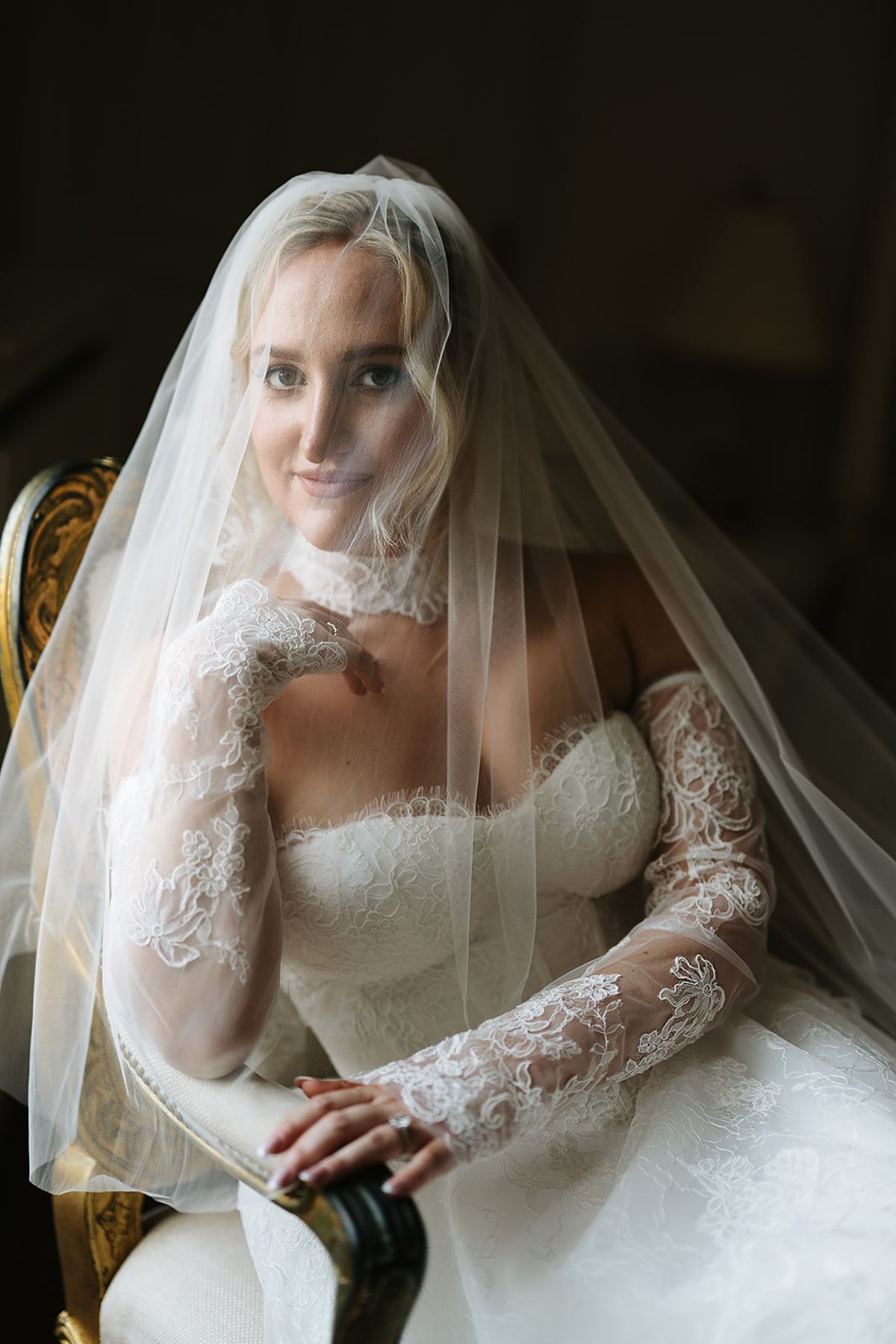 The bride sits in a gilded chair, a sheer veil over her face, wearing a lace gown with long lace sleeves and a lace collar.