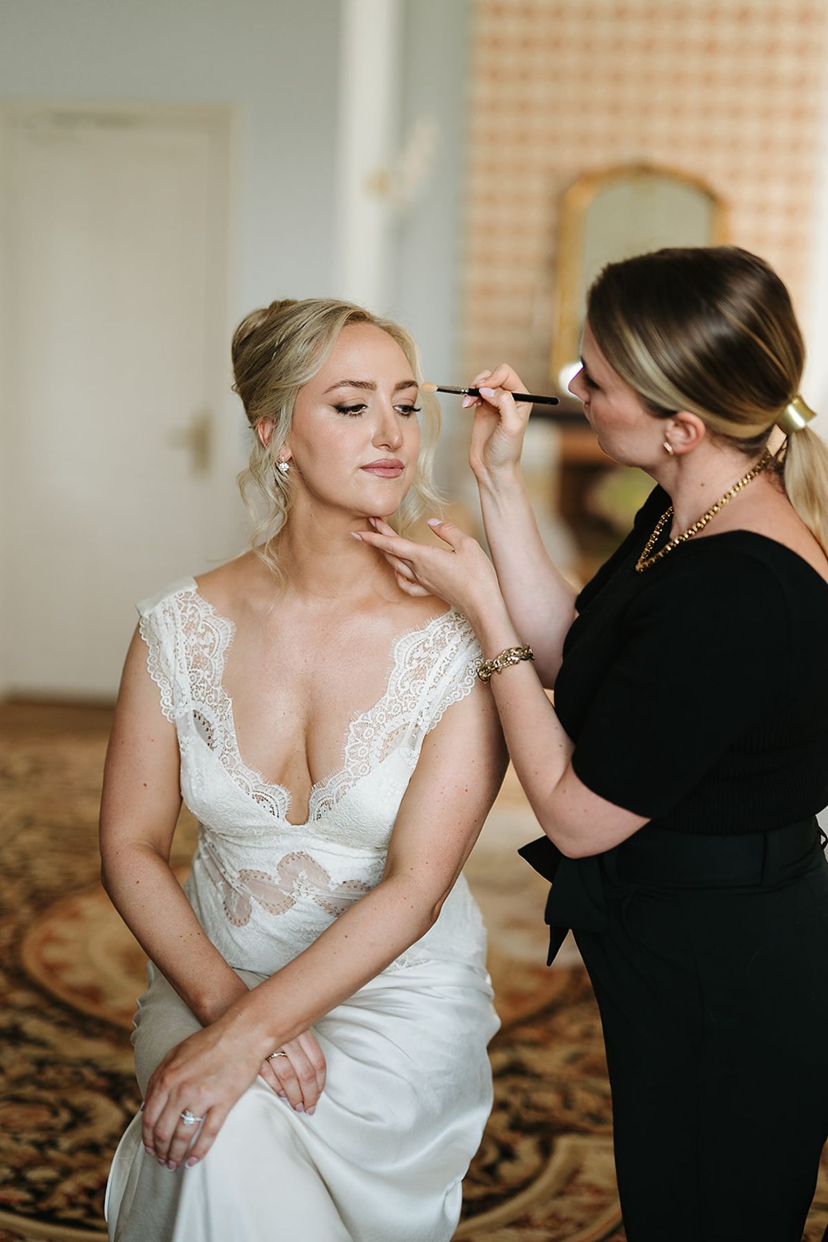 The bride, seated in a lace-trimmed dress, has make-up applied by a woman in black in an ornate room.
