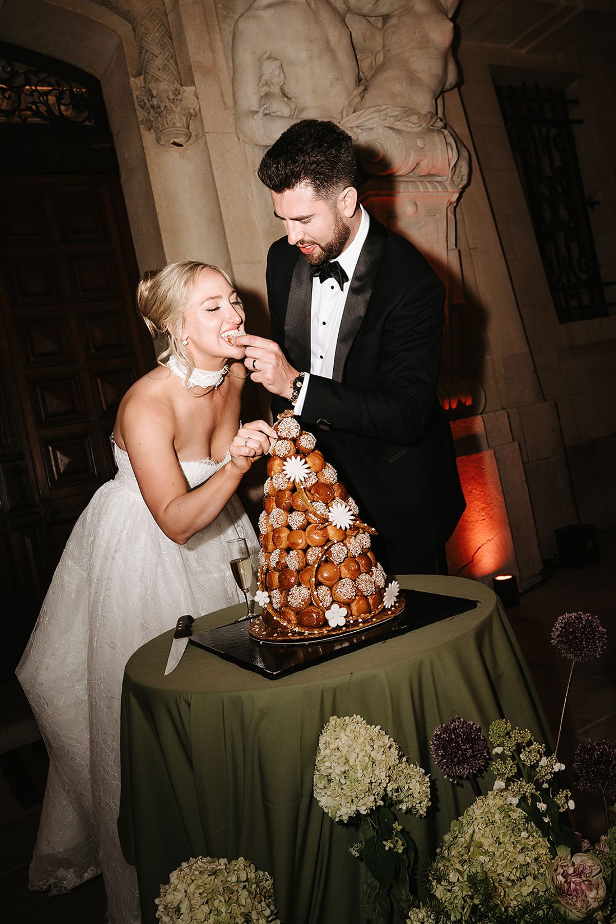 The bride and groom share a bite from a tall choux pastry tower on a table with an olive green cloth.