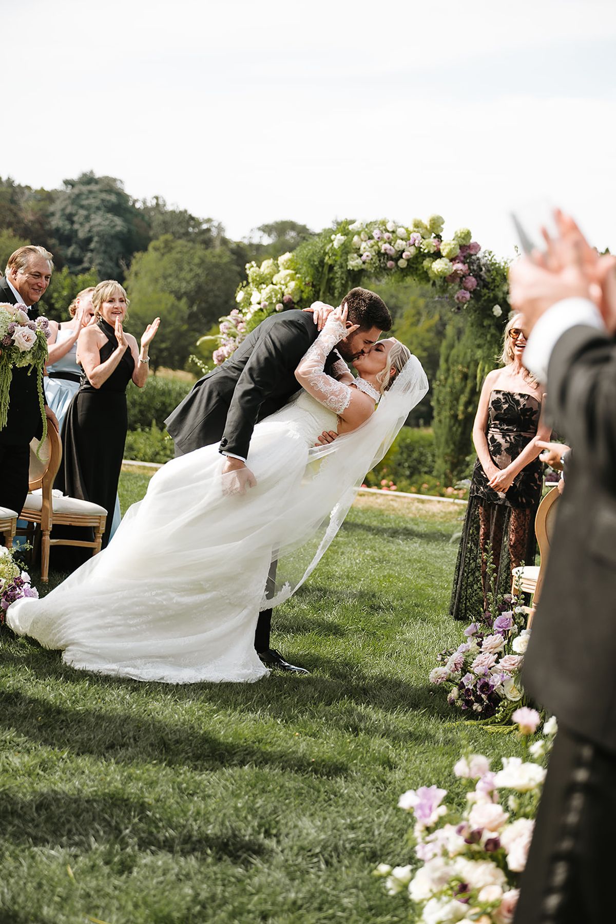 The groom dips the bride for a kiss in front of the floral arch while guests behind them applaud.