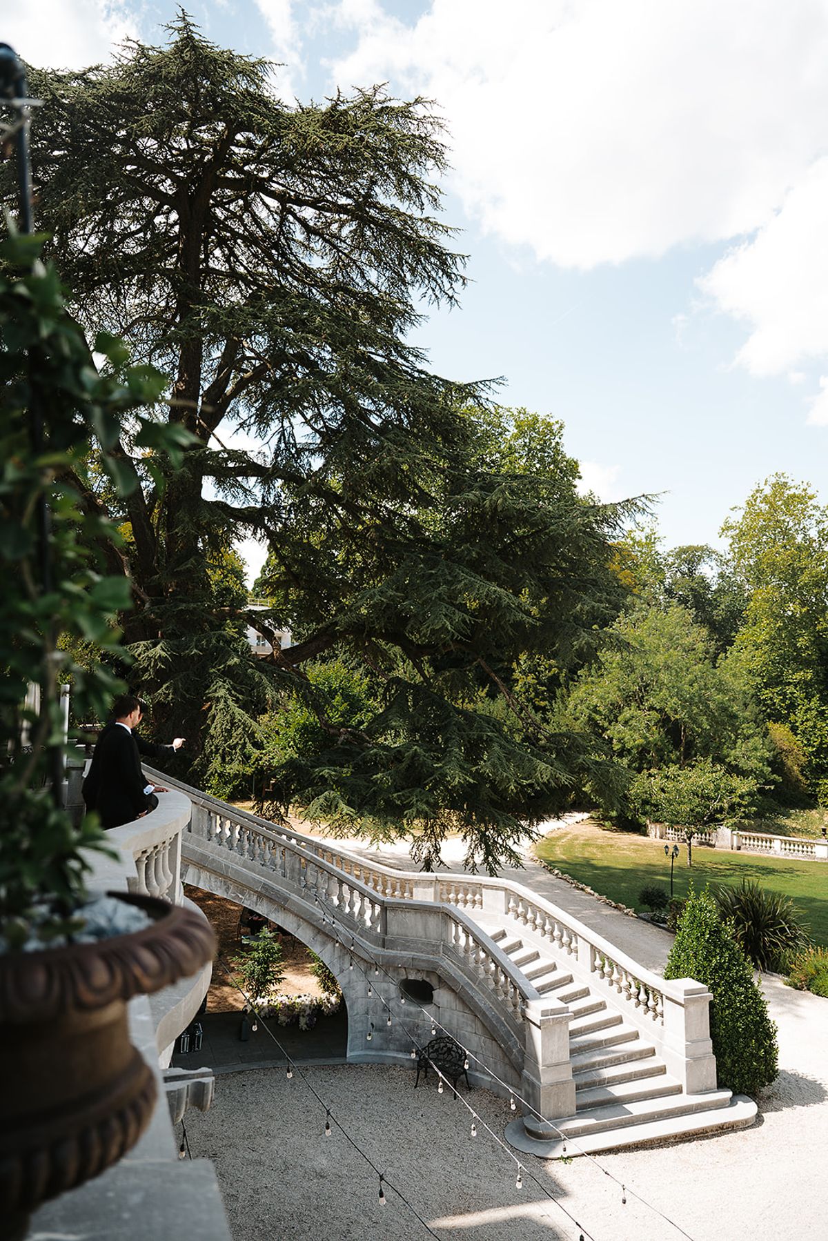 A man in dark clothing stands on a stone balcony above a curved staircase, with a large cedar tree and gardens beyond.