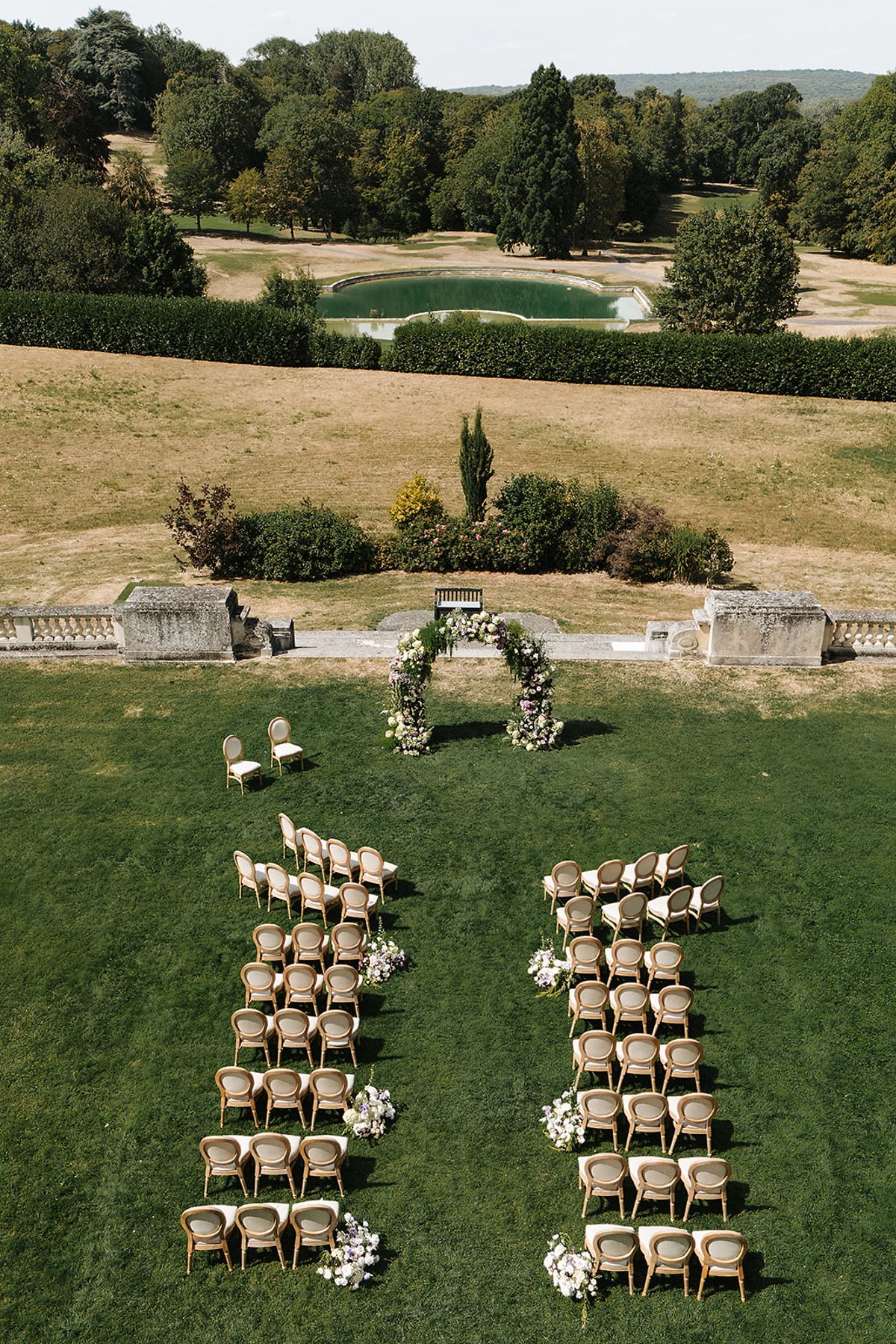 A drone view of rows of wooden chairs arranged in two blocks on a lawn facing a floral arch, with parkland beyond.