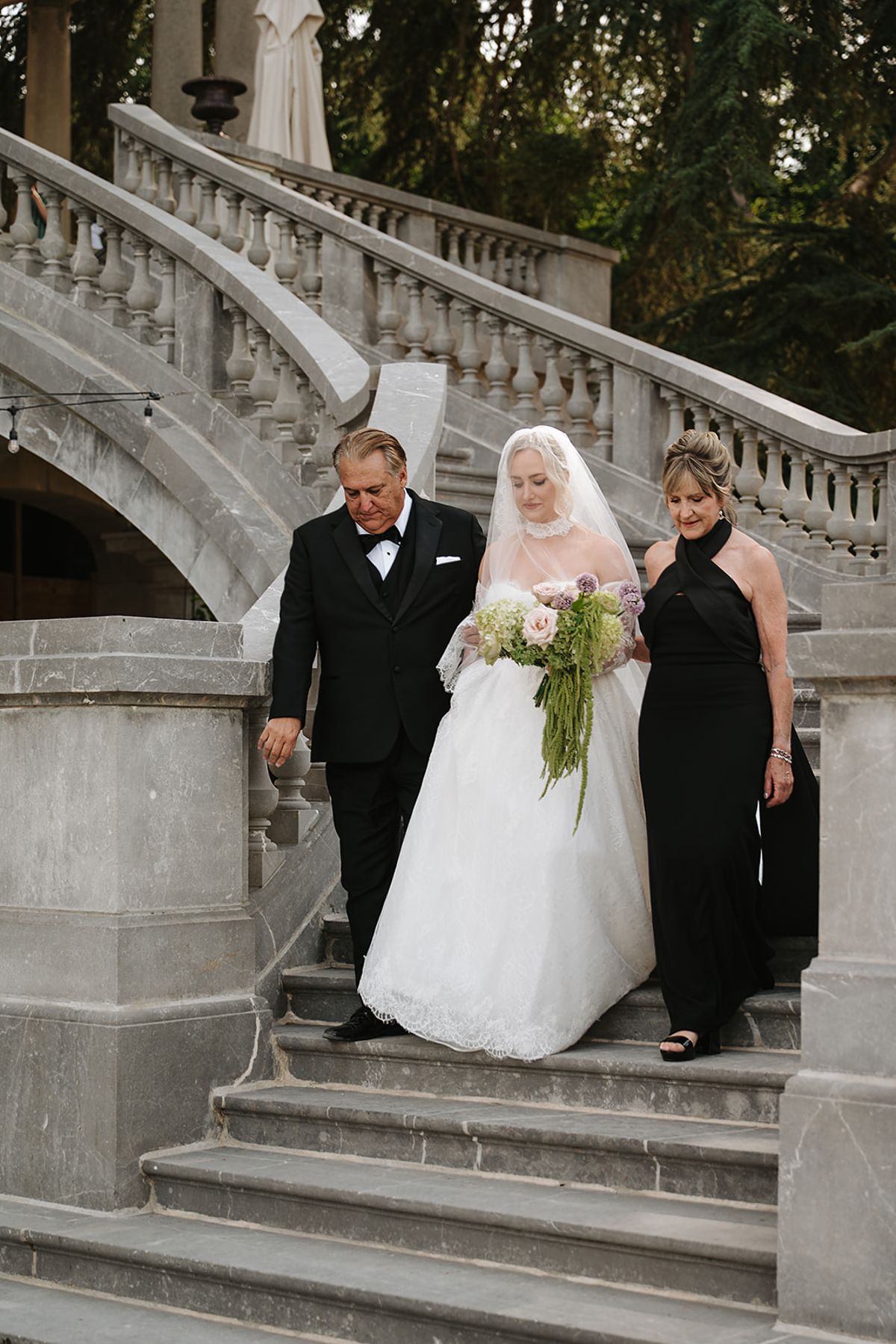 The bride in a white gown holding a trailing green bouquet descends a grand stone staircase between a man in a dark suit and a woman in black.