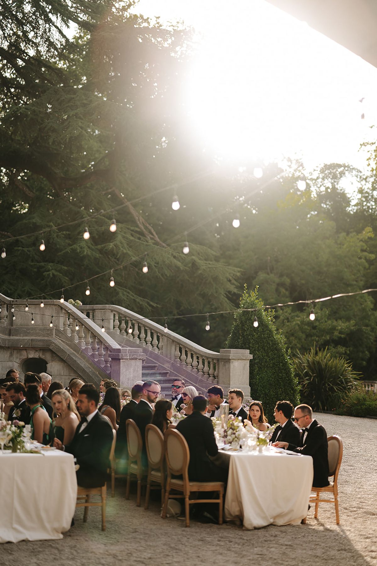 Guests in formal clothes sit along a long table beside a stone staircase, with bright sunlight and string lights through the trees.