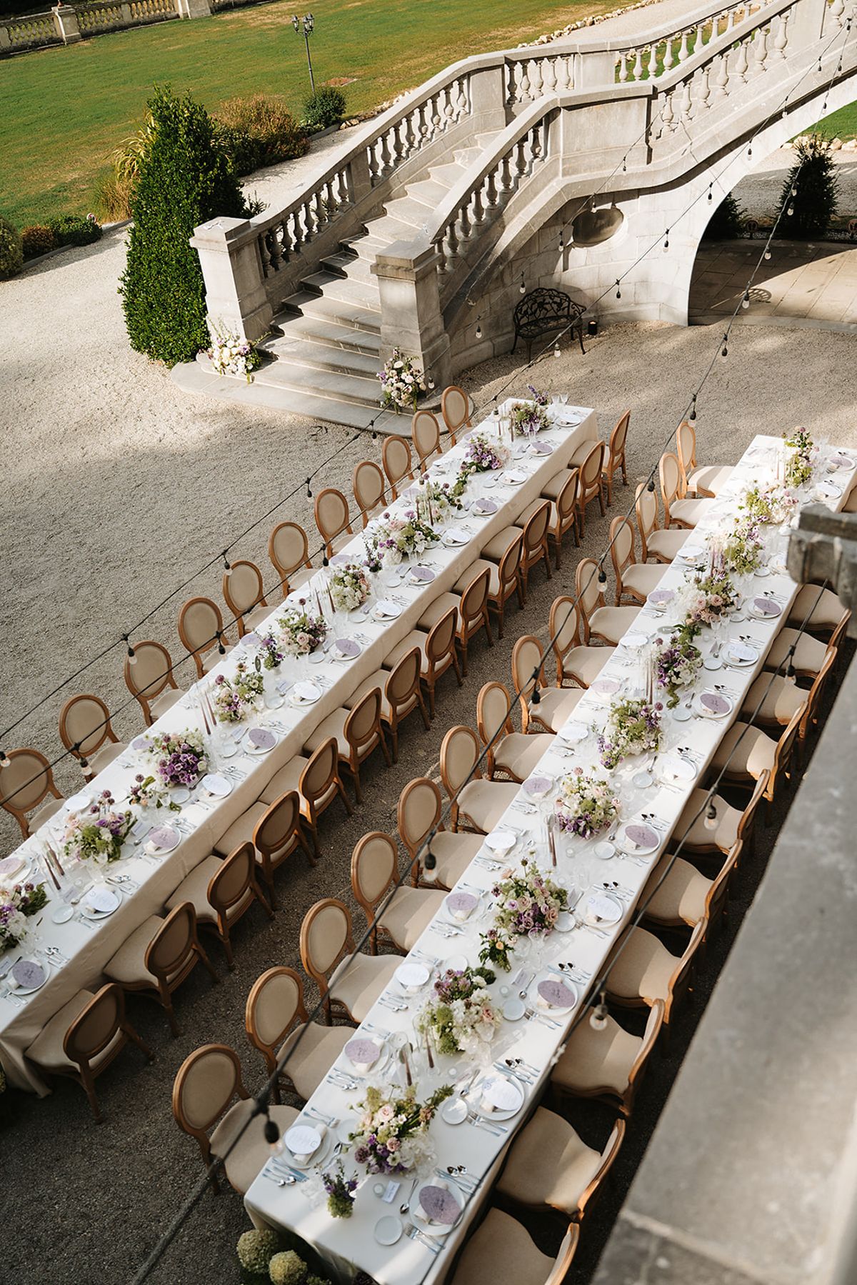 Long dining tables dressed with white linen and low flower arrangements stretch beside a sweeping stone staircase.