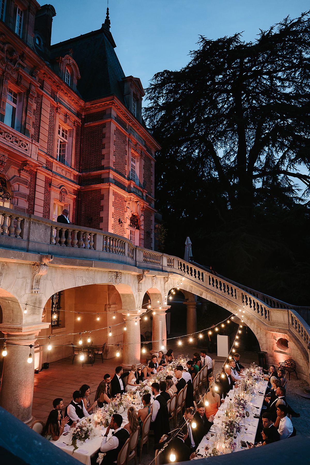 Guests sit at long tables under string lights beside a chateau lit in warm colours, with dark trees against the evening sky.
