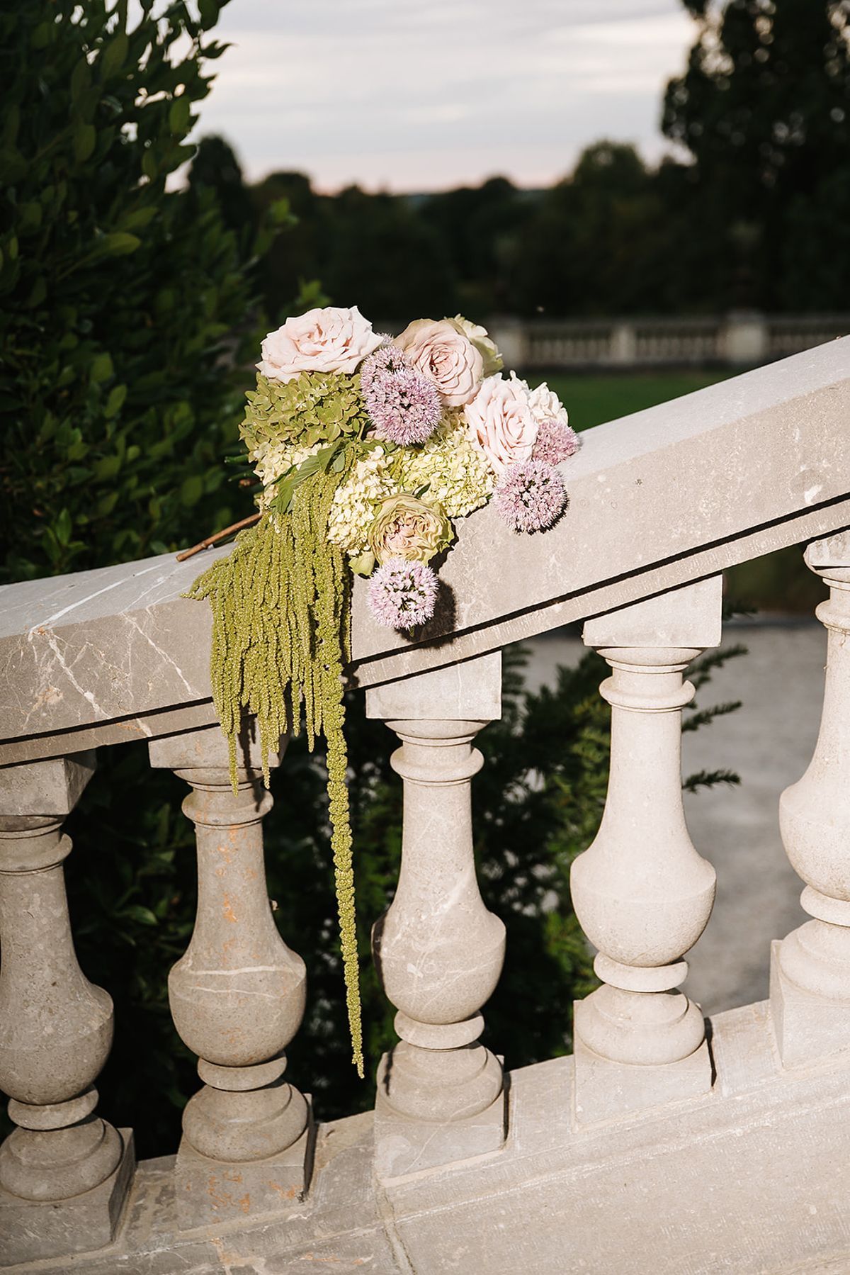 A cluster of pale pink flowers and trailing green amaranthus is fixed to a stone balustrade overlooking the grounds.