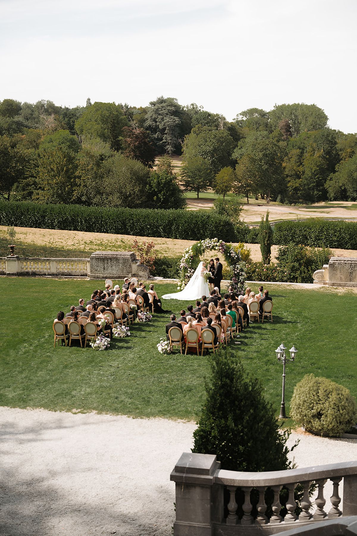 The couple stand before a floral arch facing seated guests on a lawn, with hedges, trees and a stone balustrade beyond.