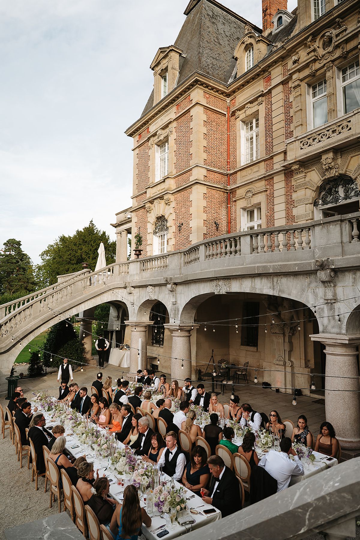 Guests sit at long dressed tables in a courtyard beneath a curved stone staircase and a brick chateau with string lights overhead.