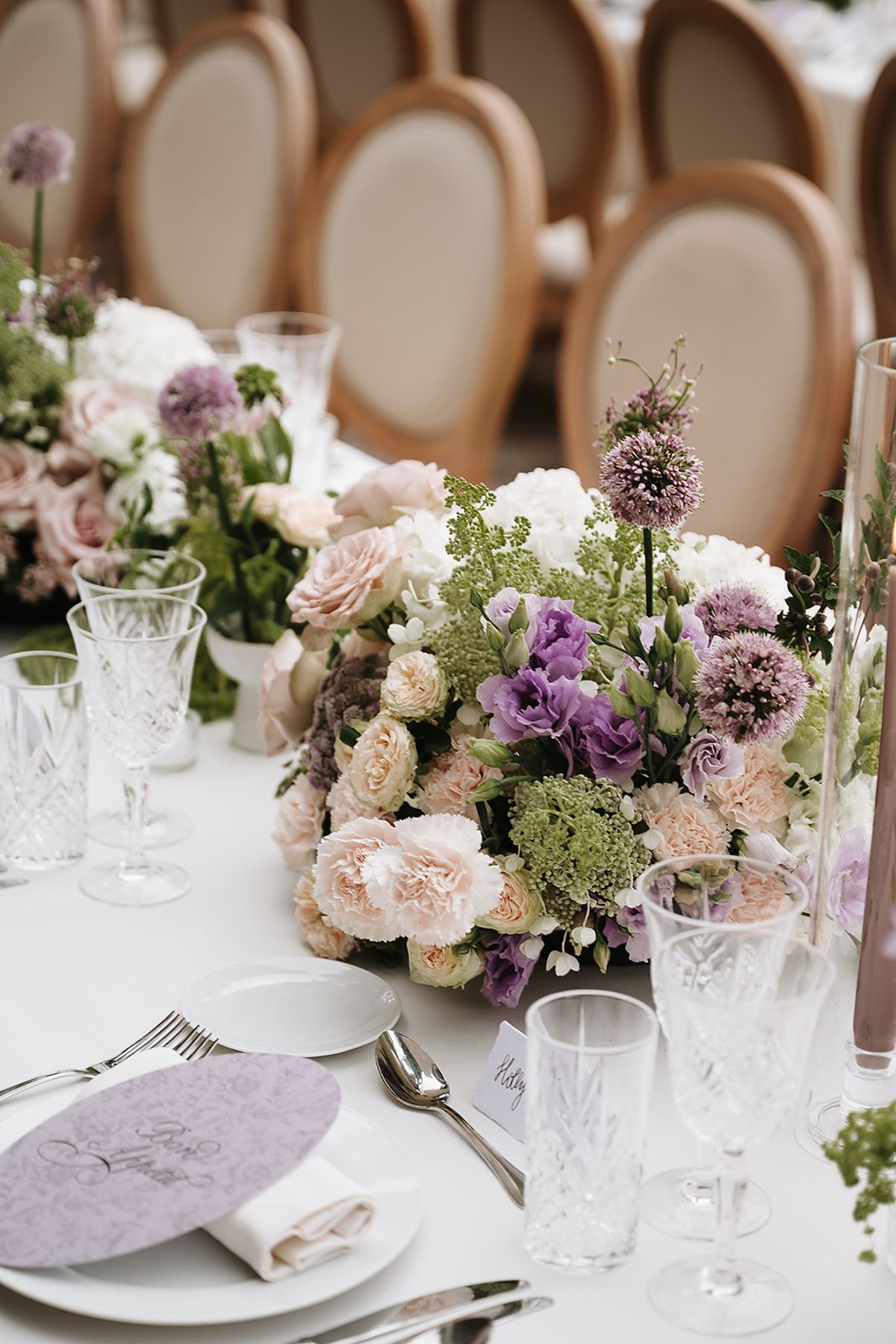 A dense centrepiece of pink, lilac and green flowers sits on a white table with crystal glasses and a lilac menu card.