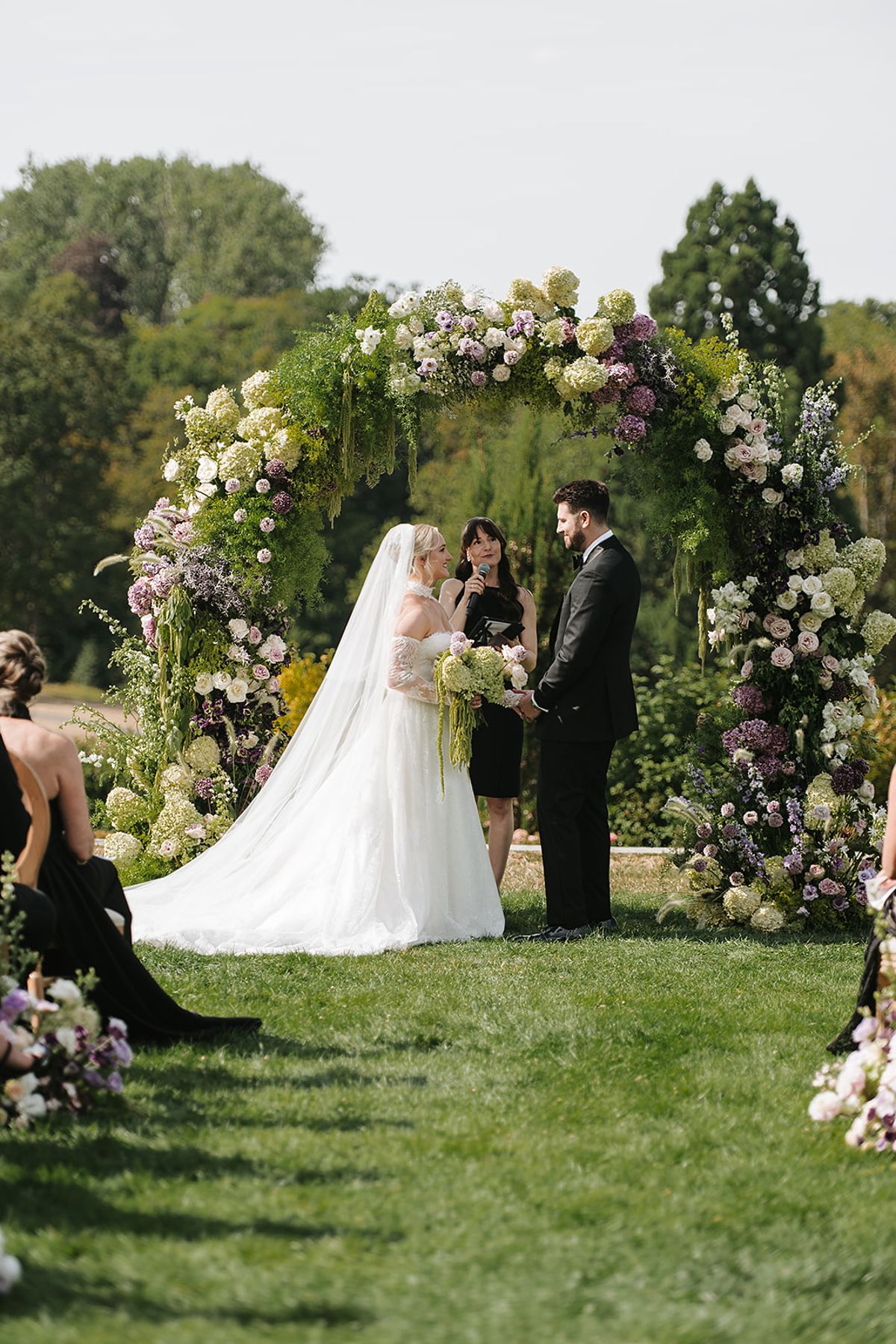 The bride in a white gown and the groom in a dark suit stand facing each other beneath a lush floral arch on a lawn.