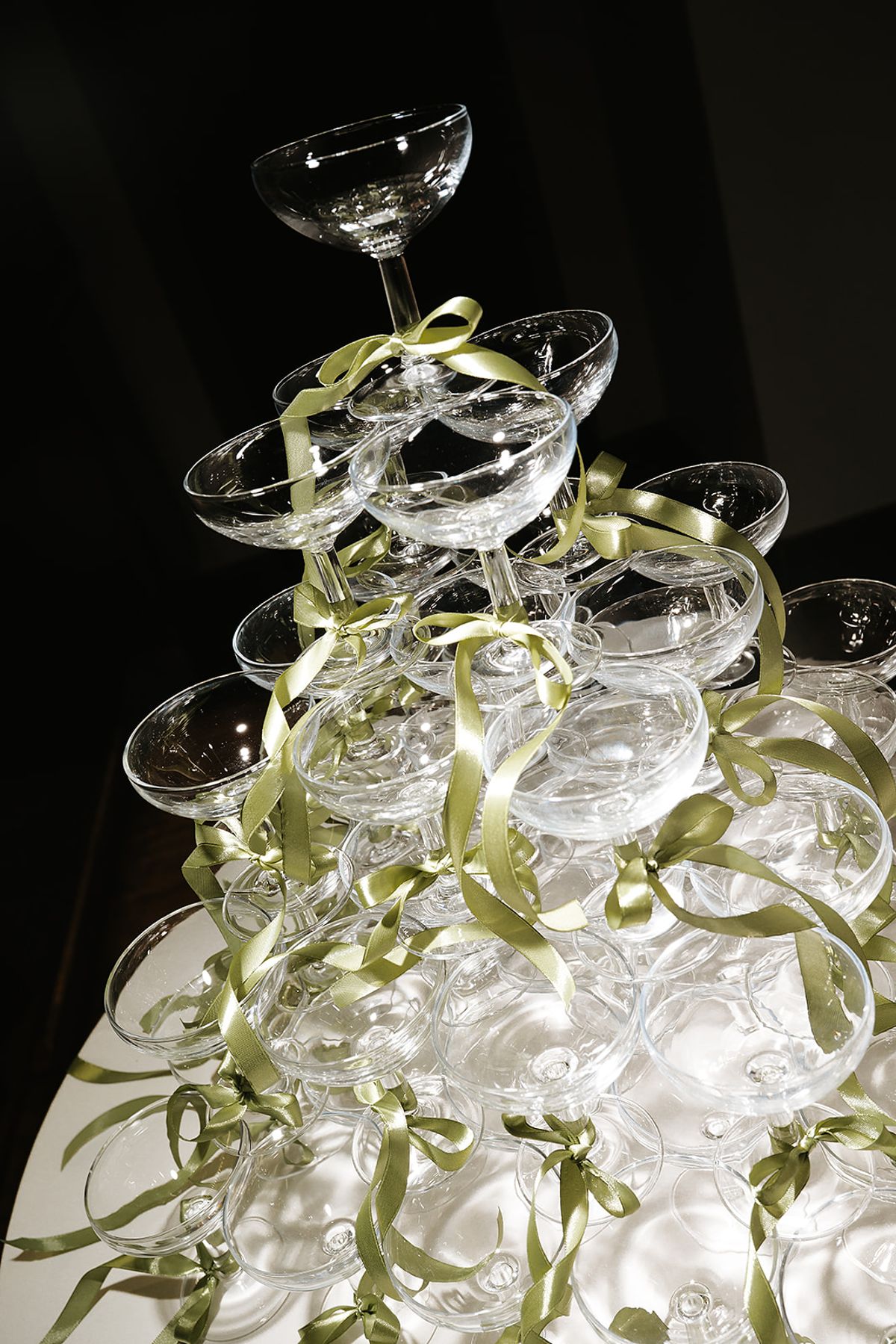 A stacked tower of champagne coupes is decorated with trailing green ribbons against a dark background.