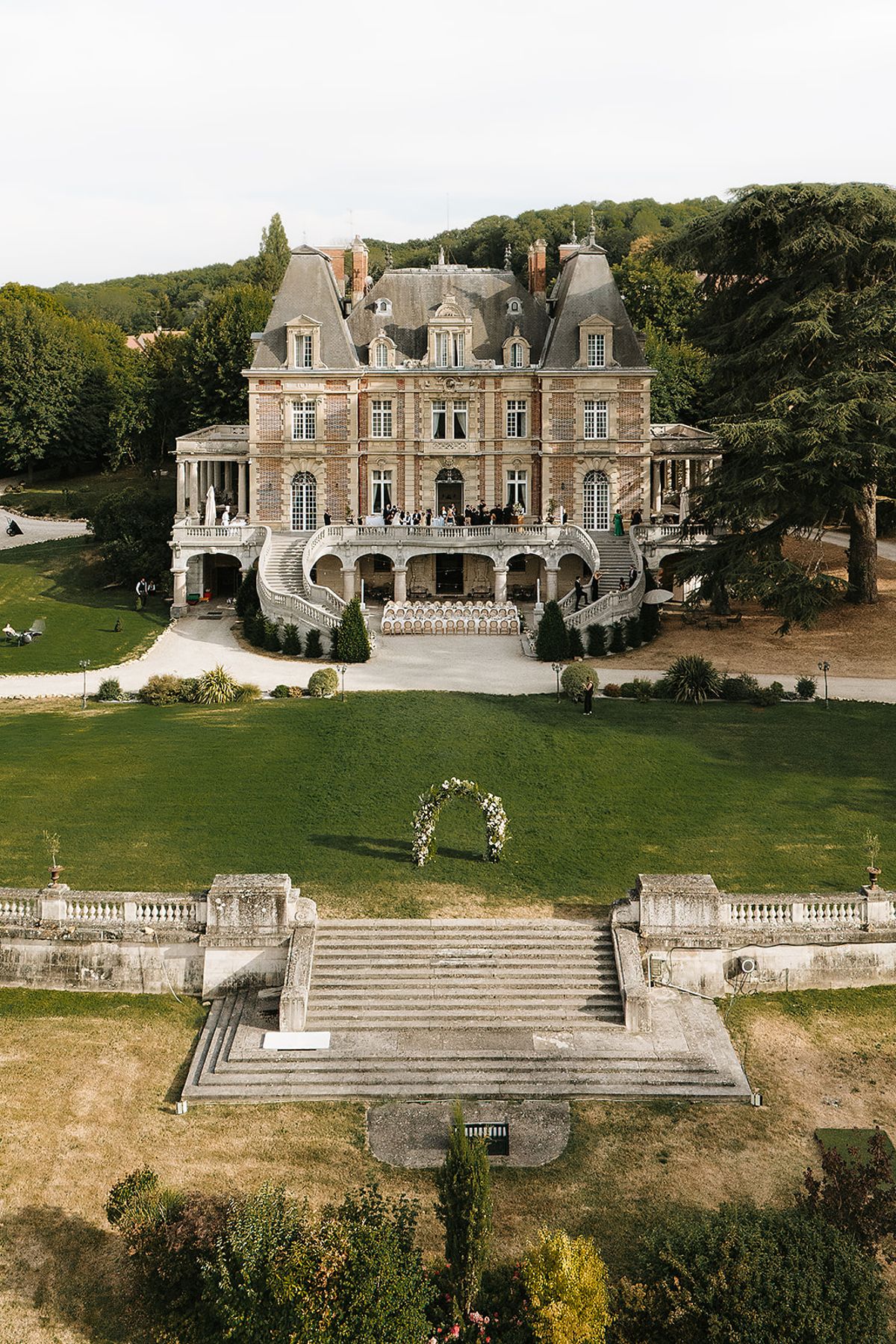 A drone view of a stone chateau with a wide lawn, stone staircase and a small floral arch set on the grass.