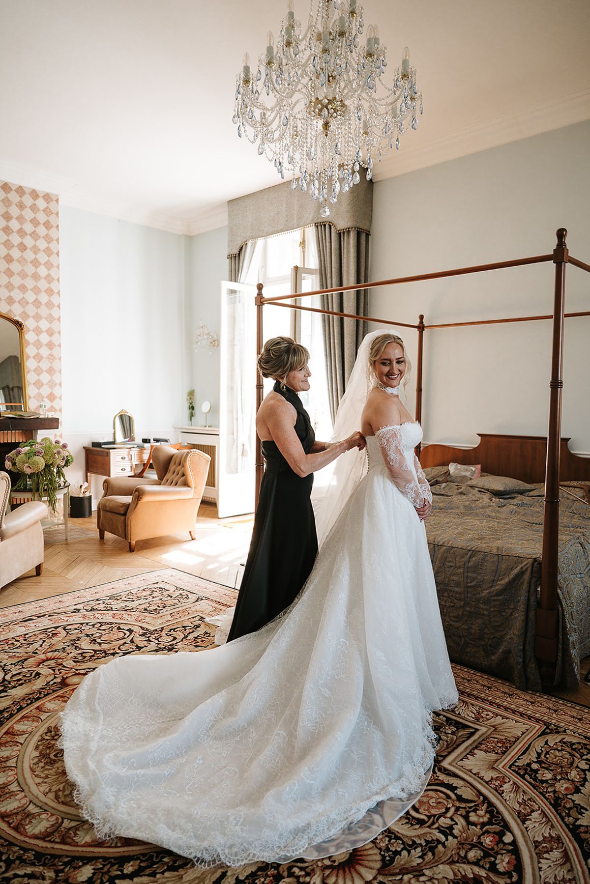 The bride in a long lace gown stands in a bedroom with a chandelier while a woman in black adjusts the back.