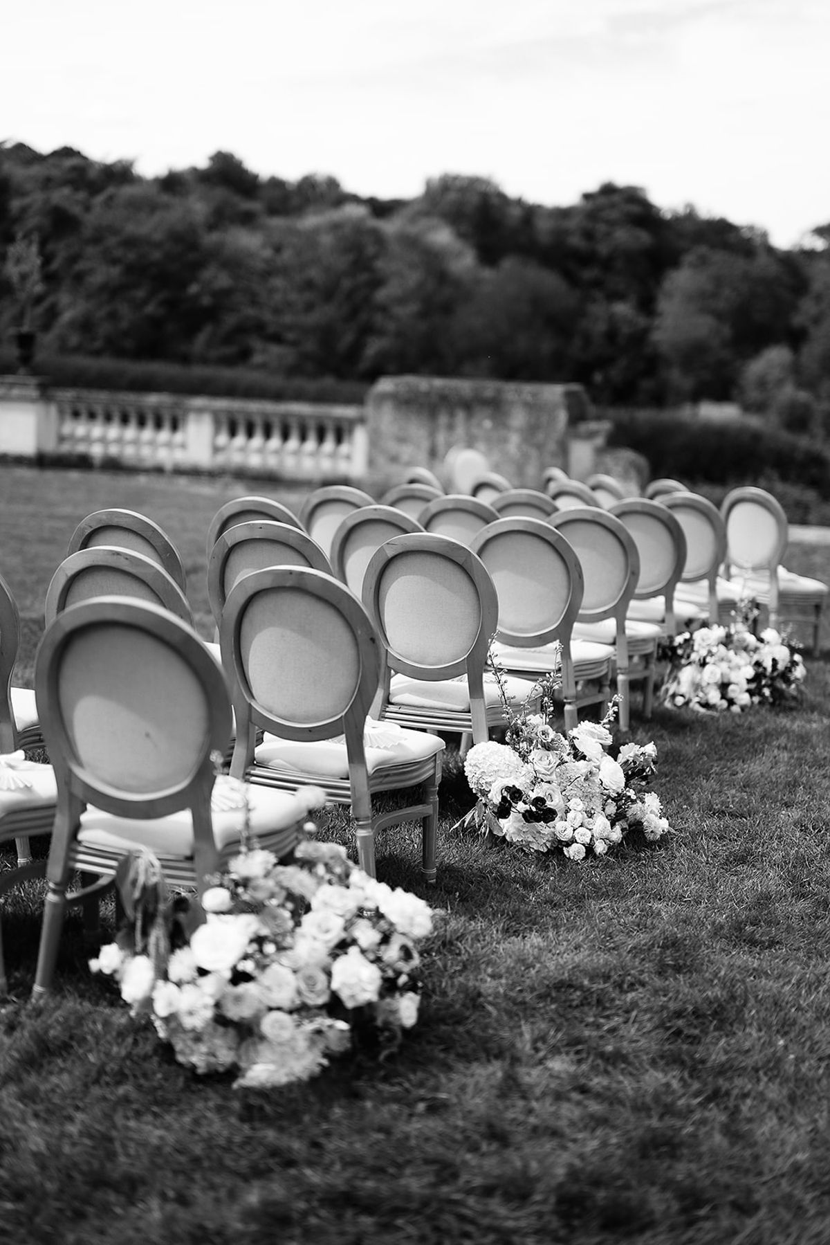 In black and white, rows of round-backed chairs line a grass aisle with clusters of flowers placed along the ground.