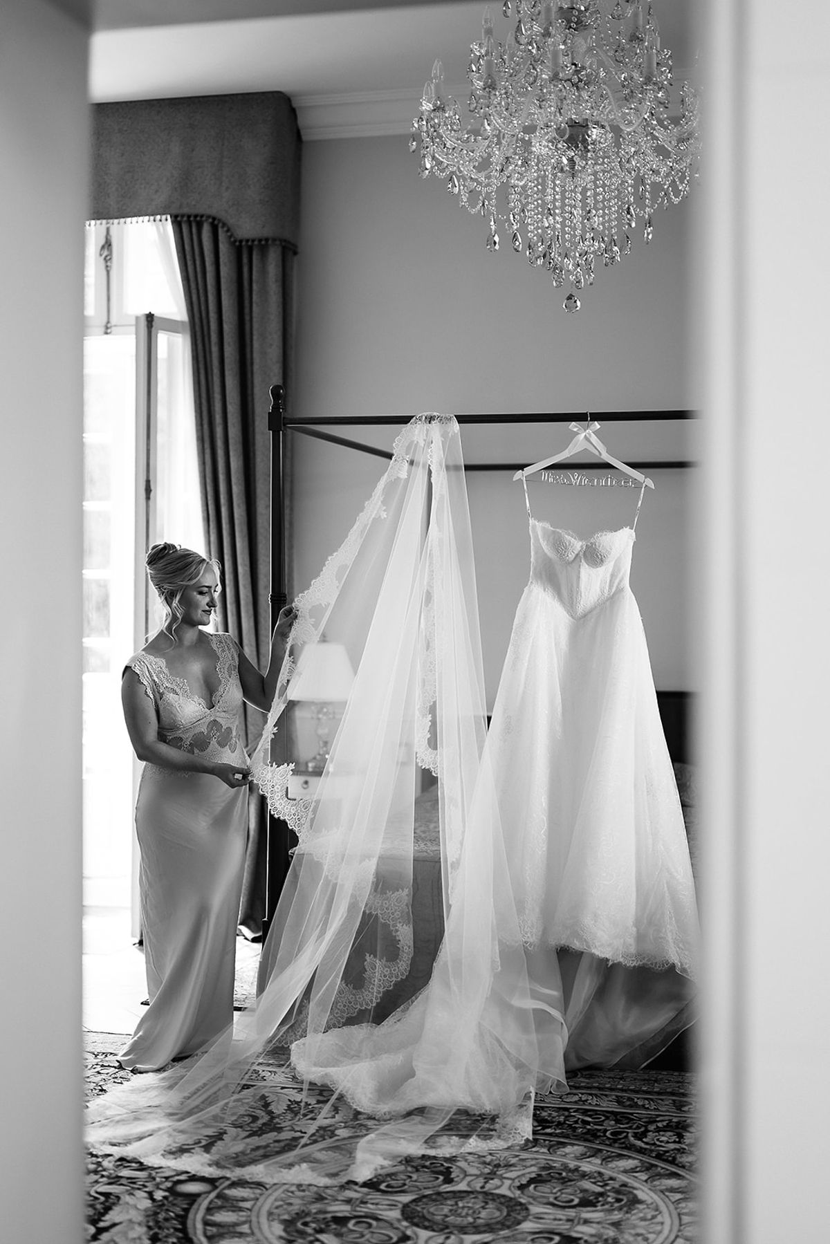 In black and white, a long veil and wedding dress hang near a window and chandelier while a woman adjusts the veil.