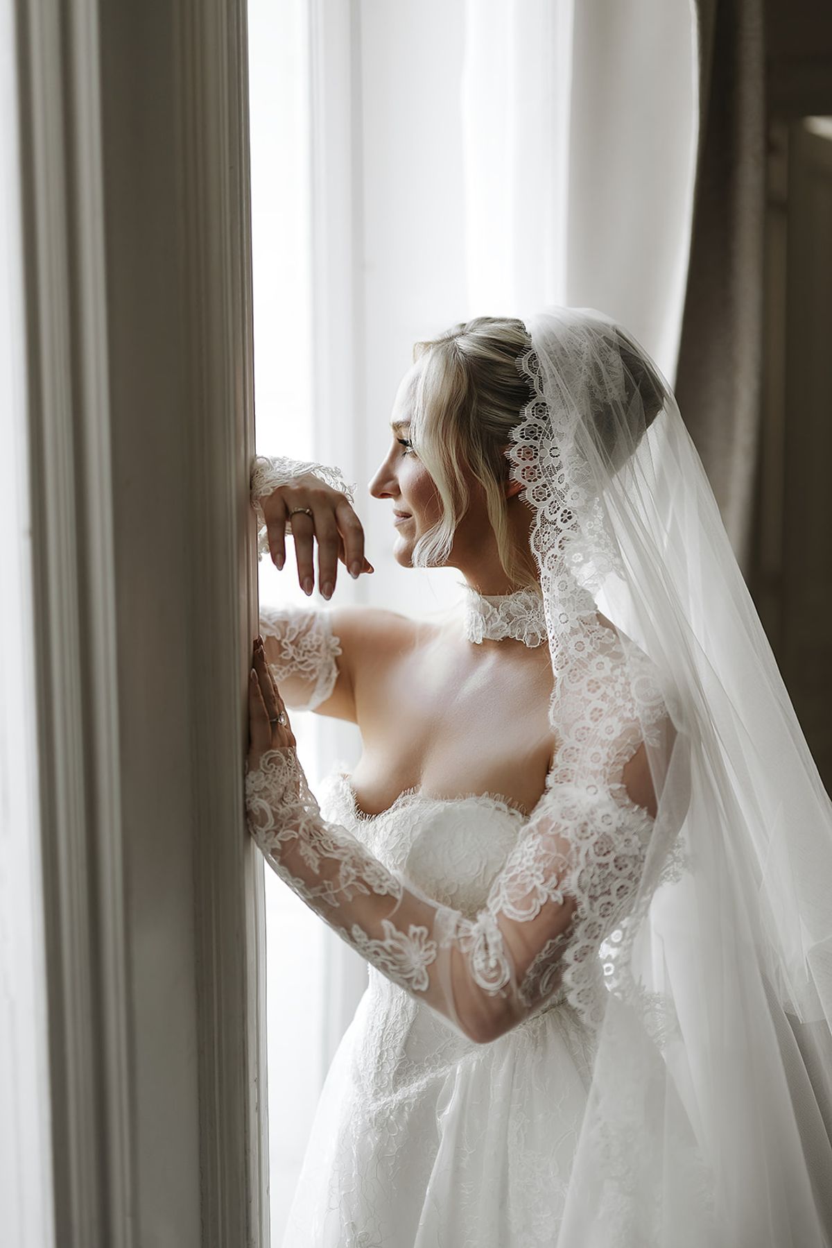 The bride in a veil and lace-sleeved gown leans against a tall window frame and smiles in soft light.