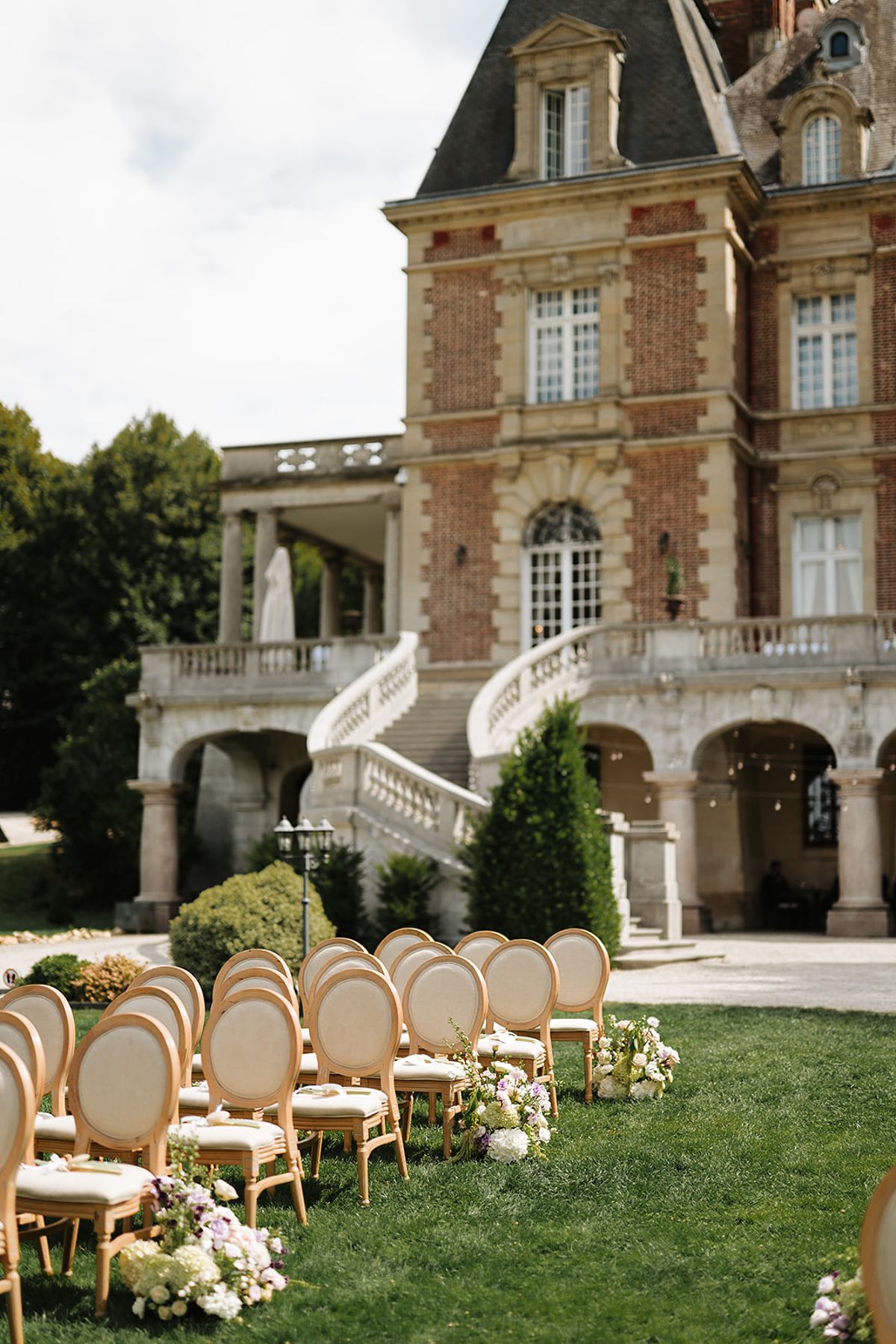 Wooden chairs are set out on a lawn in front of a red brick chateau with a stone terrace and staircase.