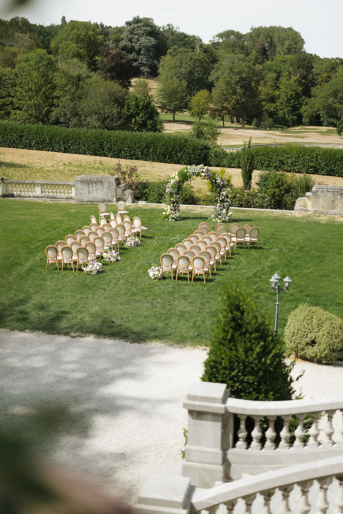 Two blocks of wooden chairs face a floral arch on a green lawn, seen from a stone terrace with wooded parkland beyond.