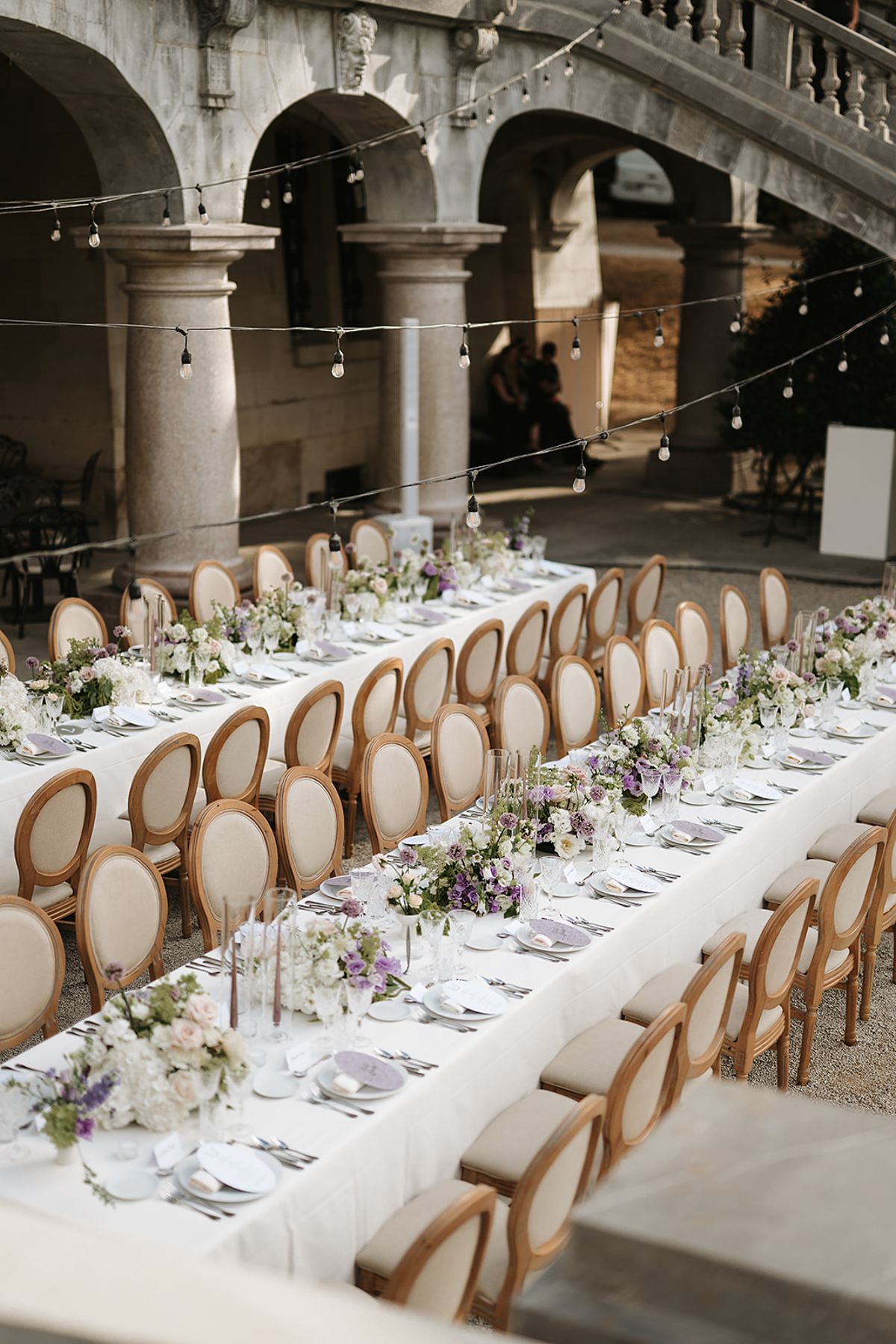 A long table with white cloth, flowers and round-backed chairs stretches along a stone arcade hung with string lights.