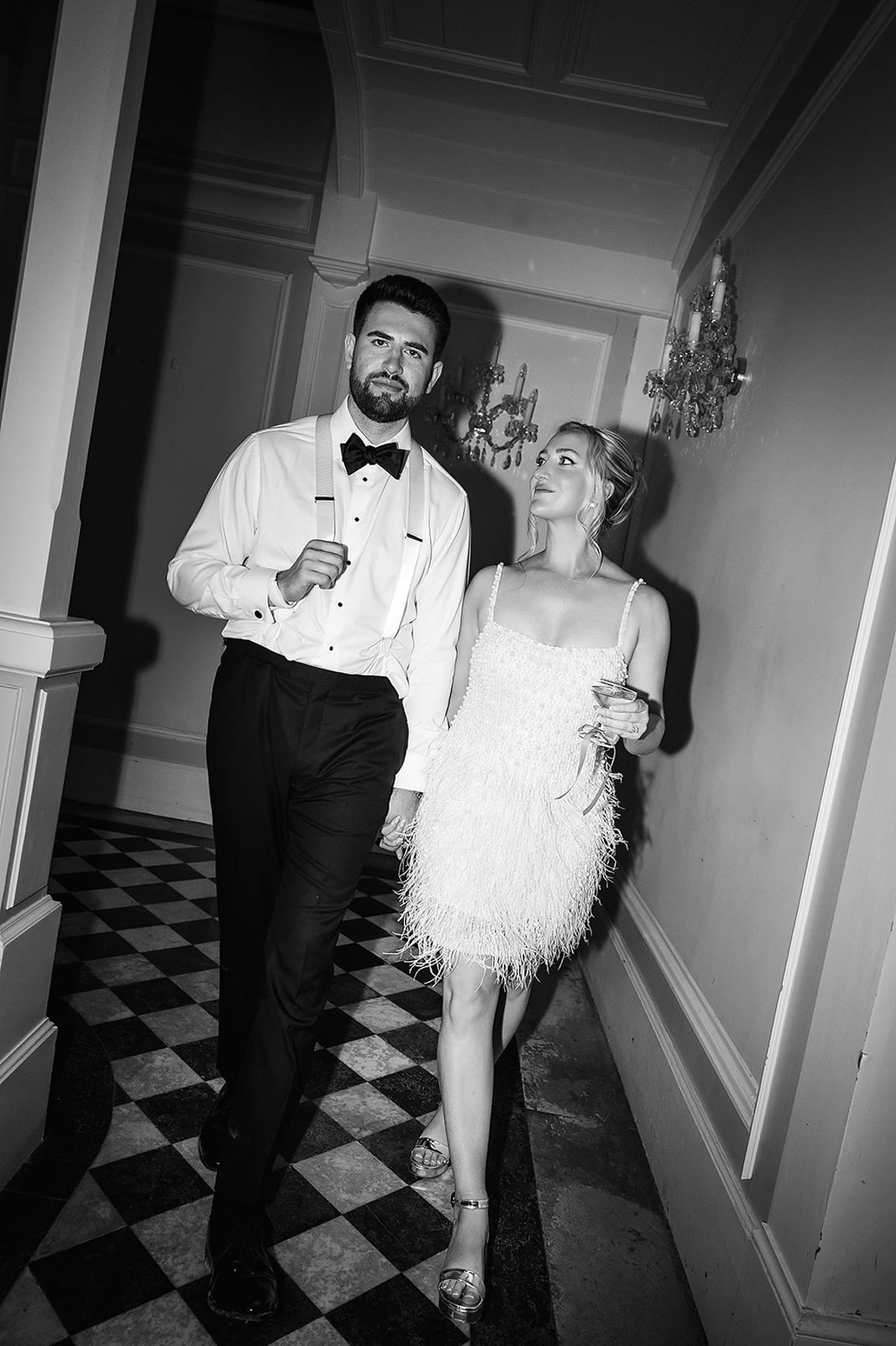 In black and white, the groom in a bow tie and the bride in a short feathered dress walk along a chequered-floor corridor.