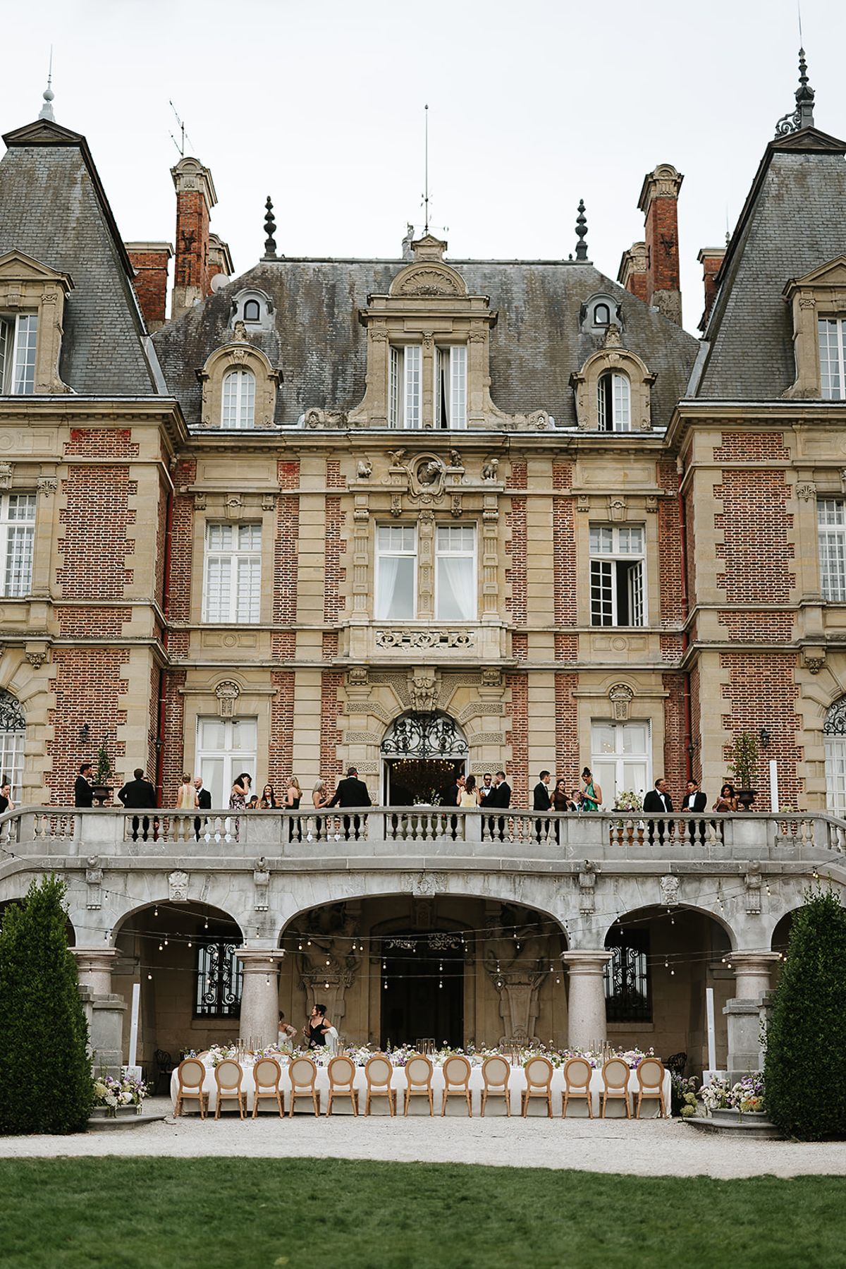 A grand red brick and stone chateau with slate roofs, a balcony lined with guests and a long table beneath.