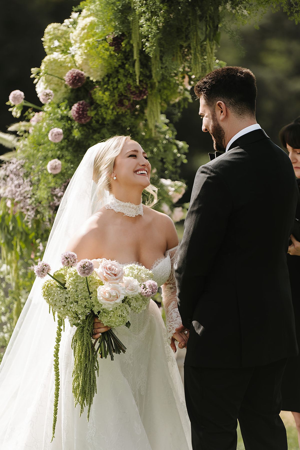 The smiling bride in a veil and lace gown holds a pale bouquet and faces the groom in a black suit, before a floral arch.