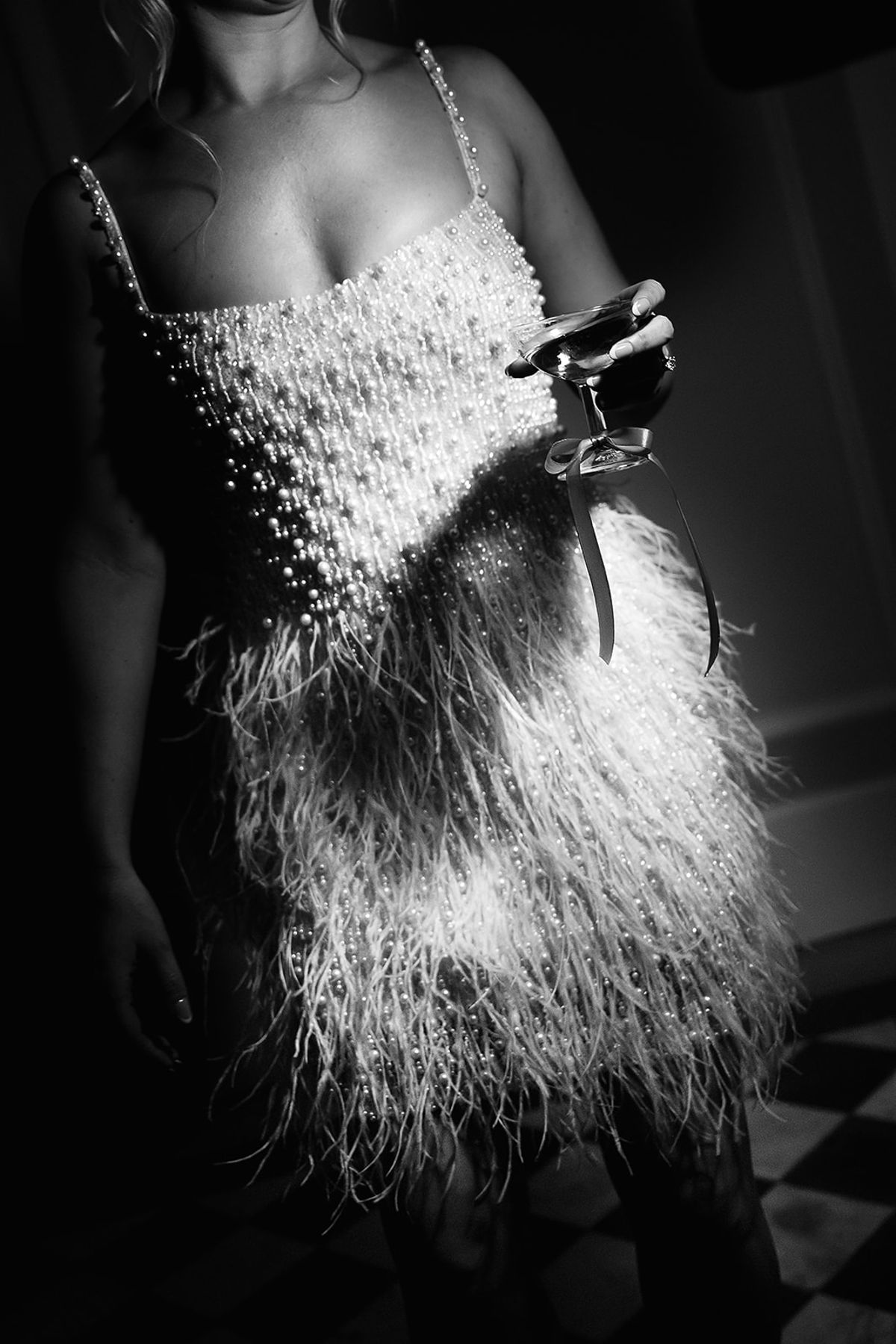 Black and white shot of a woman in a pearl-beaded feather mini dress, holding a coupe glass with a ribbon on its stem.