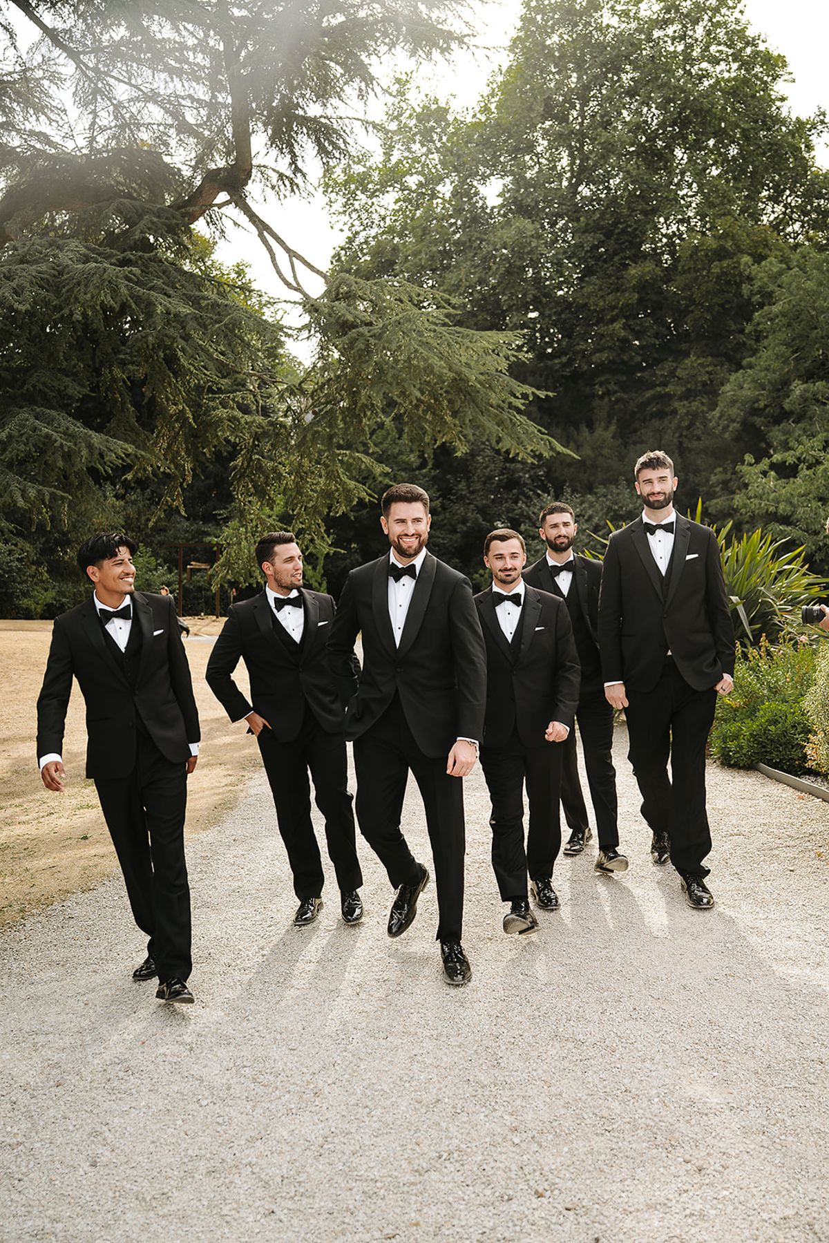 Seven men in black suits and bow ties walk together towards the camera along a gravel path lined with tall trees.