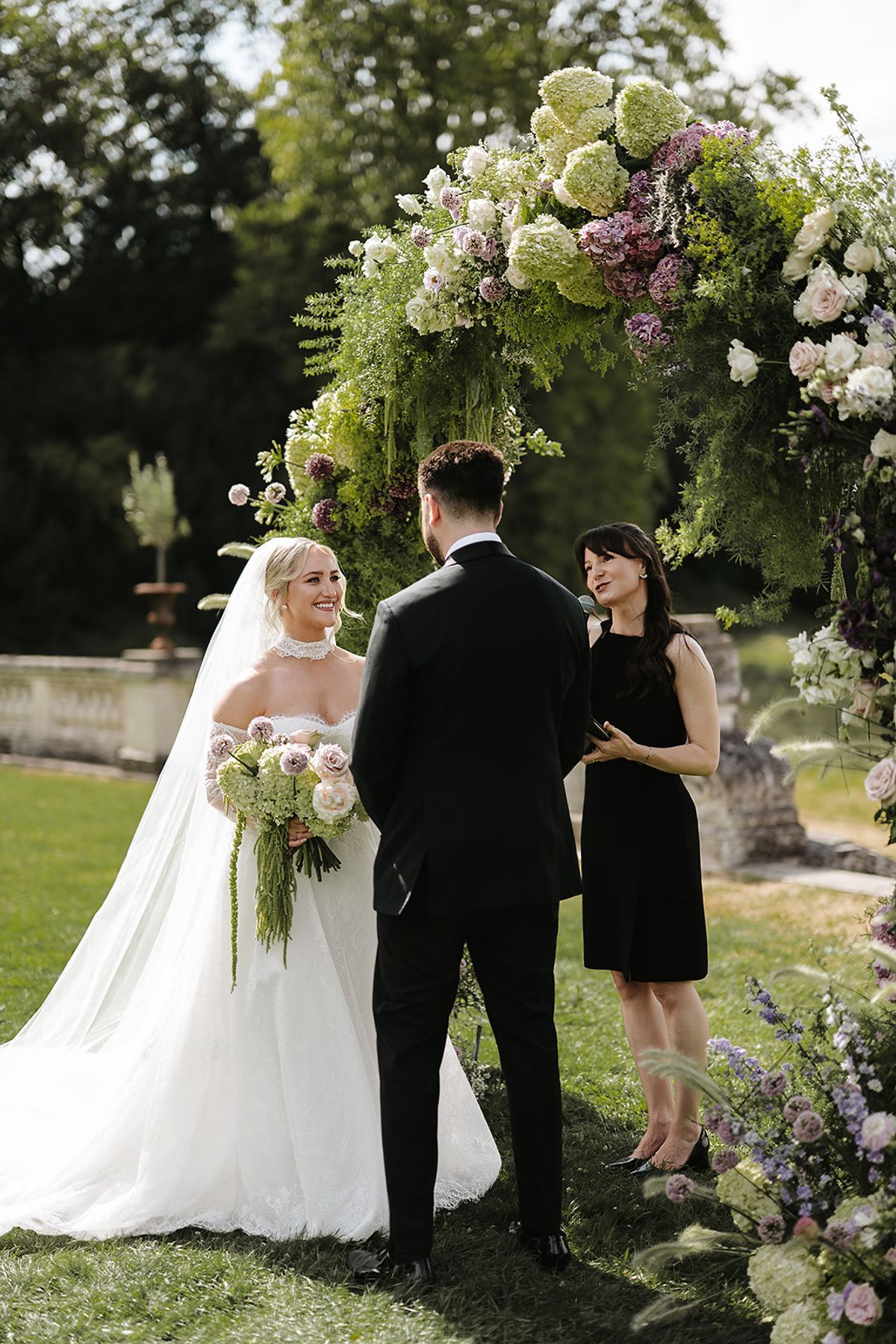 The bride in a white gown and the groom in a black suit stand beneath a flower arch beside a woman in a black dress.