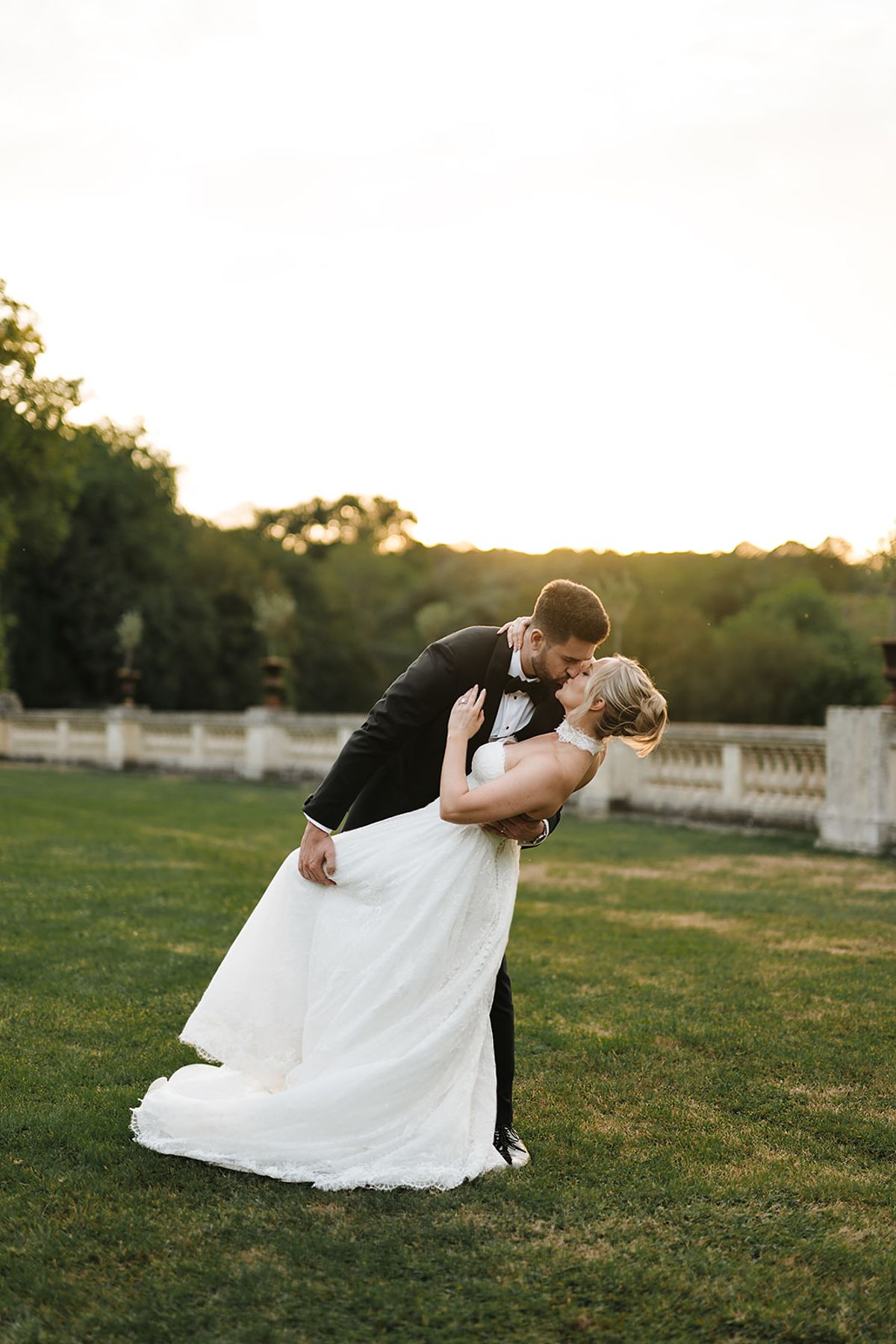 The couple share a dipped kiss on a lawn, the bride's long white skirt flowing, with wooded hills glowing in low light behind.