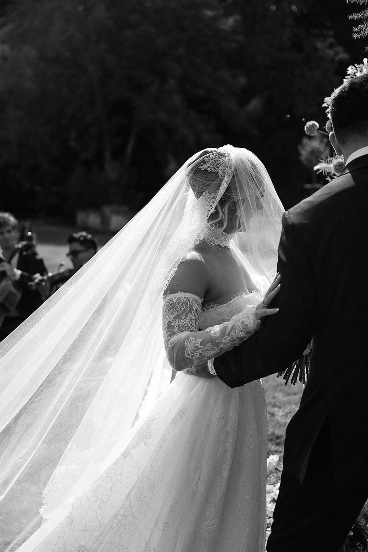 The bride in a lace gown and long veil rests her hand on the groom's arm, while guests watch outdoors in black and white.
