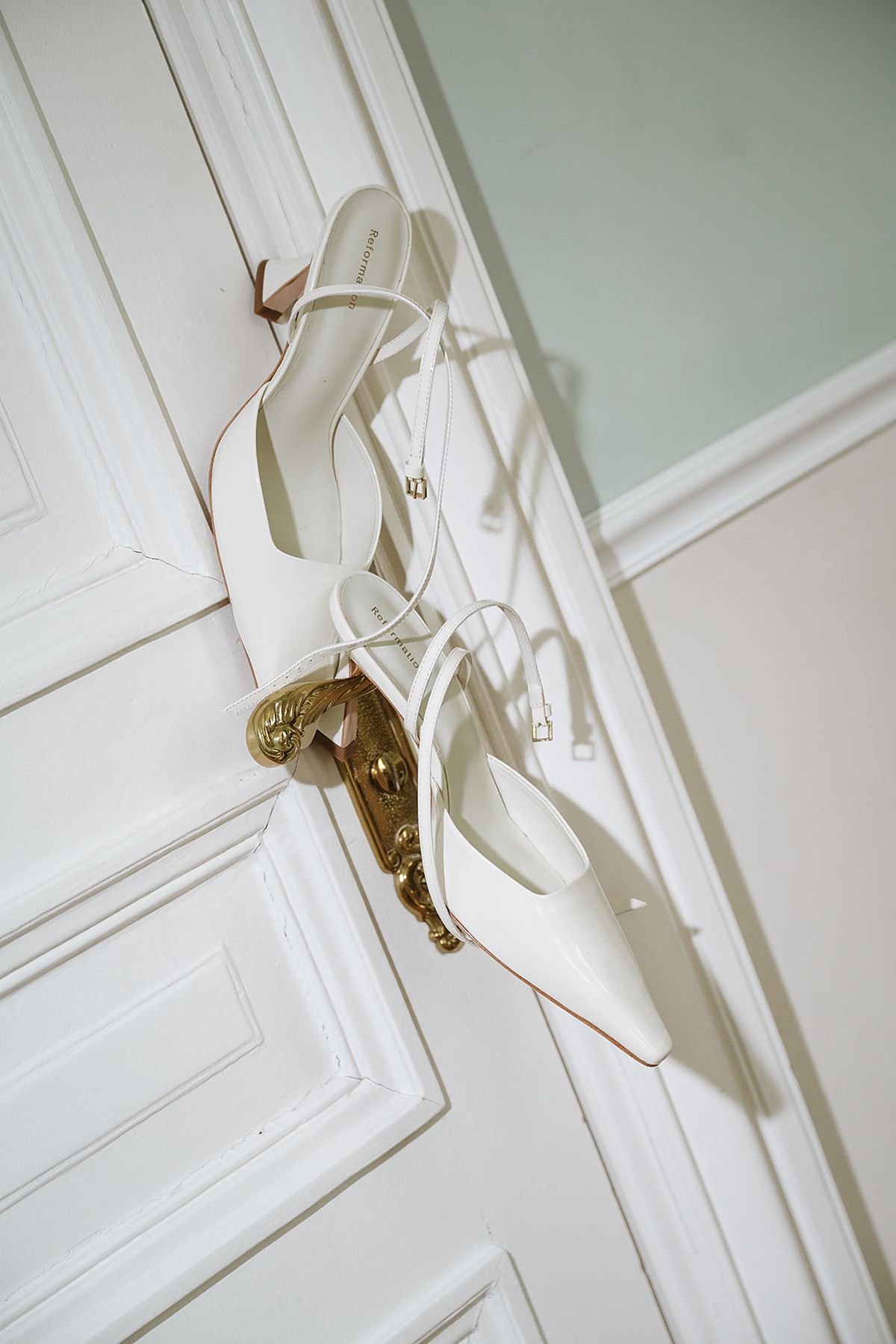 A pair of white pointed slingback heels with thin ankle straps hangs from an ornate brass handle on a white panelled door.