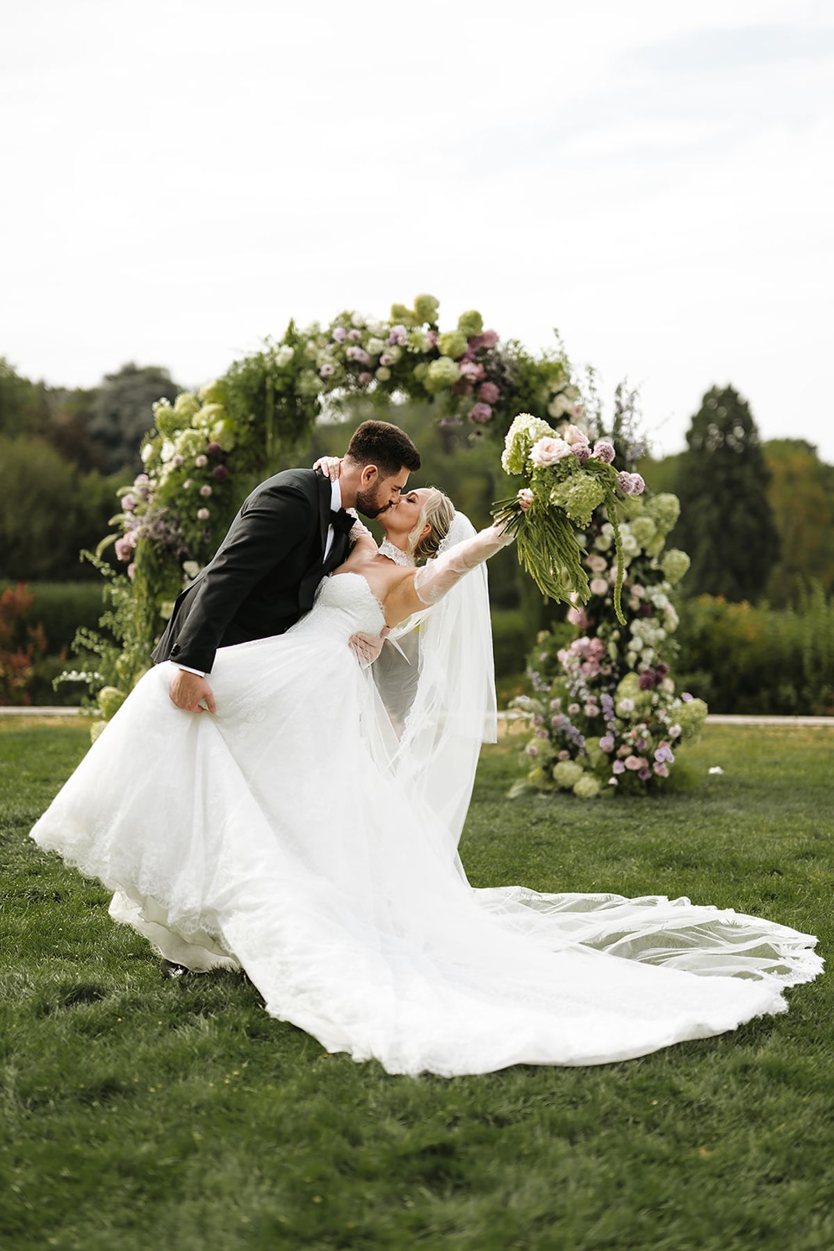 The groom leans the bride back in a kiss in front of a floral arch, her veil and train trailing across the grass.