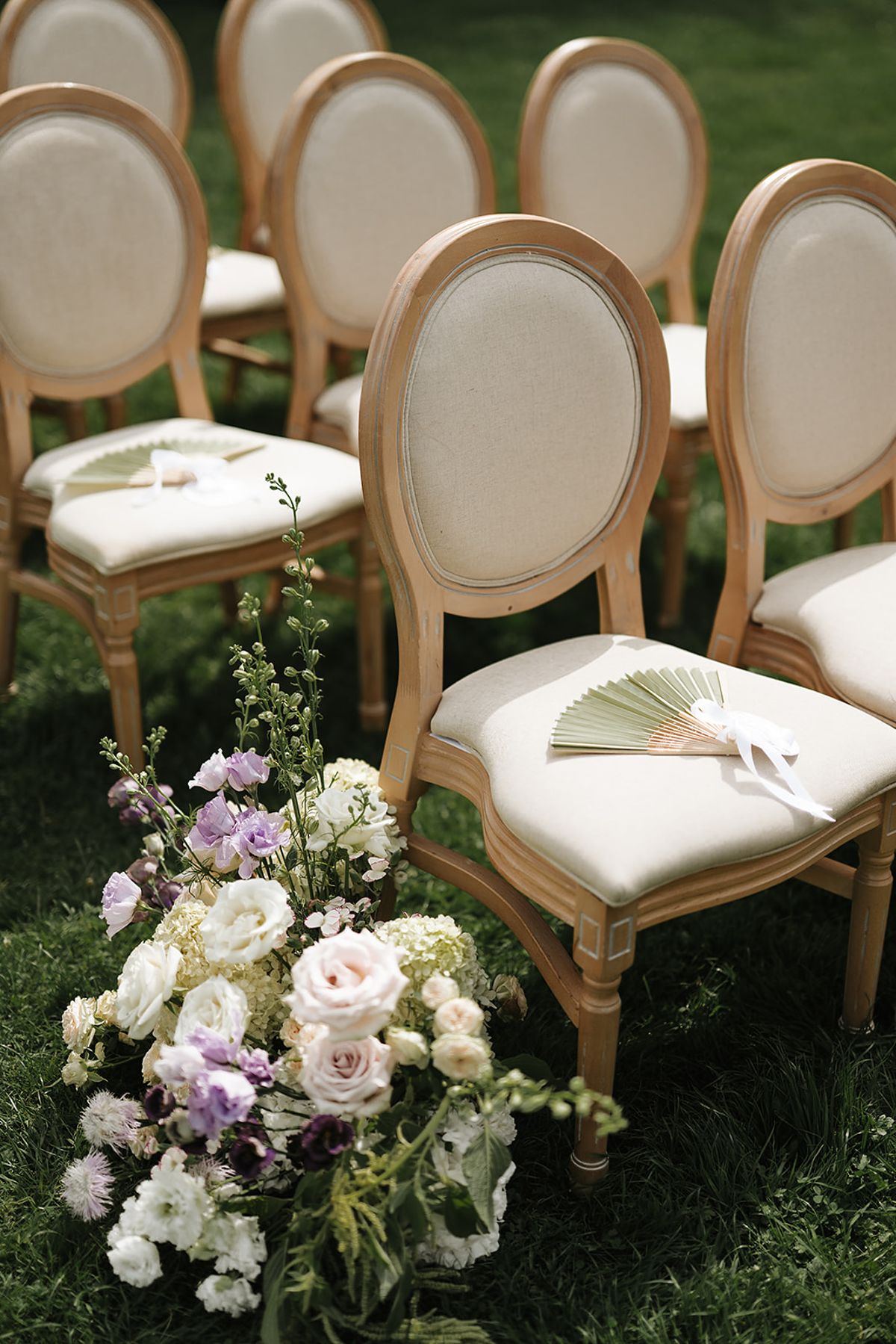 Rows of cream and wooden chairs on grass, each with a green fan, beside a bouquet of white and lilac flowers.