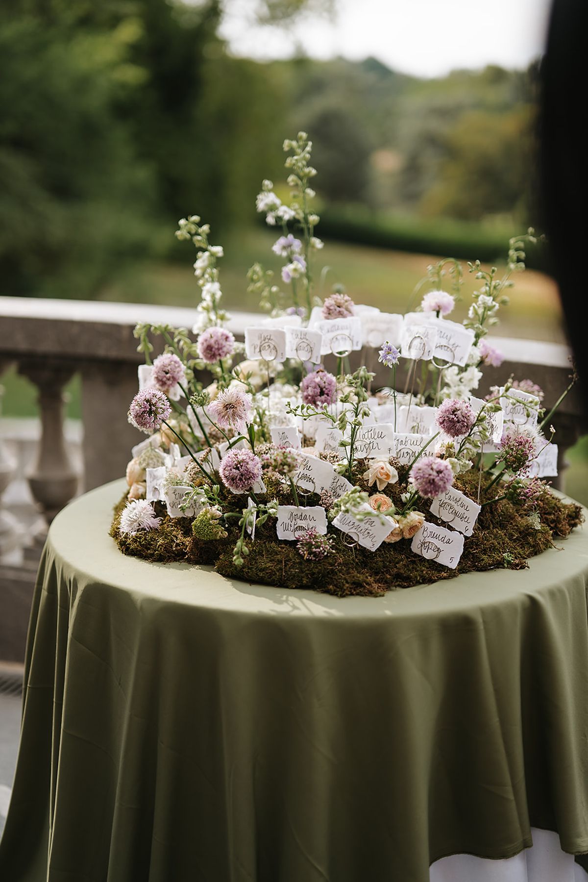 A round table with an olive green cloth holds a low arrangement of pink and lilac flowers and moss with small place cards, on a terrace.