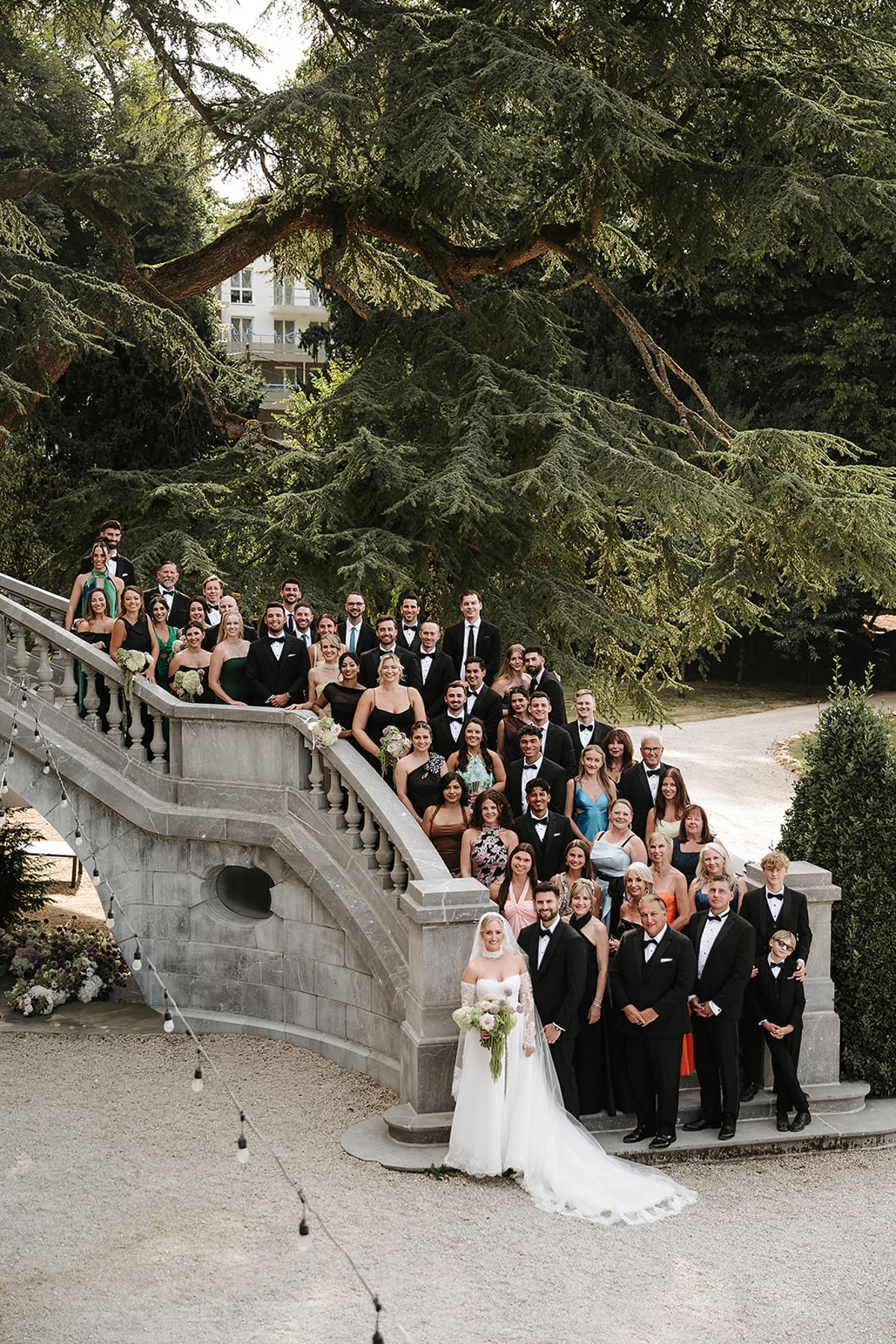 A large group of guests dressed mostly in black pose on a stone staircase, with the bride and groom at the bottom.