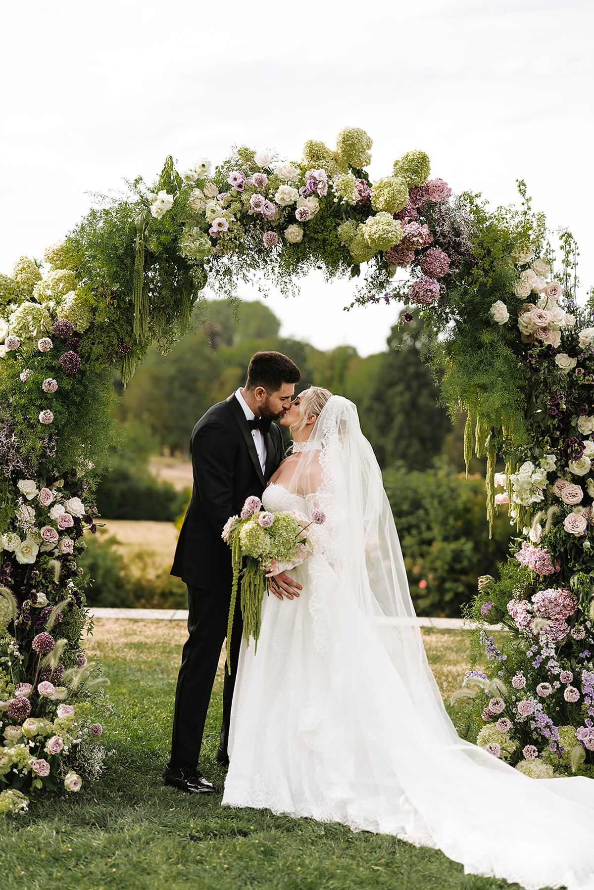 The bride in a white veiled gown and the groom in a black tuxedo kiss beneath a round arch of pink, mauve and green flowers.