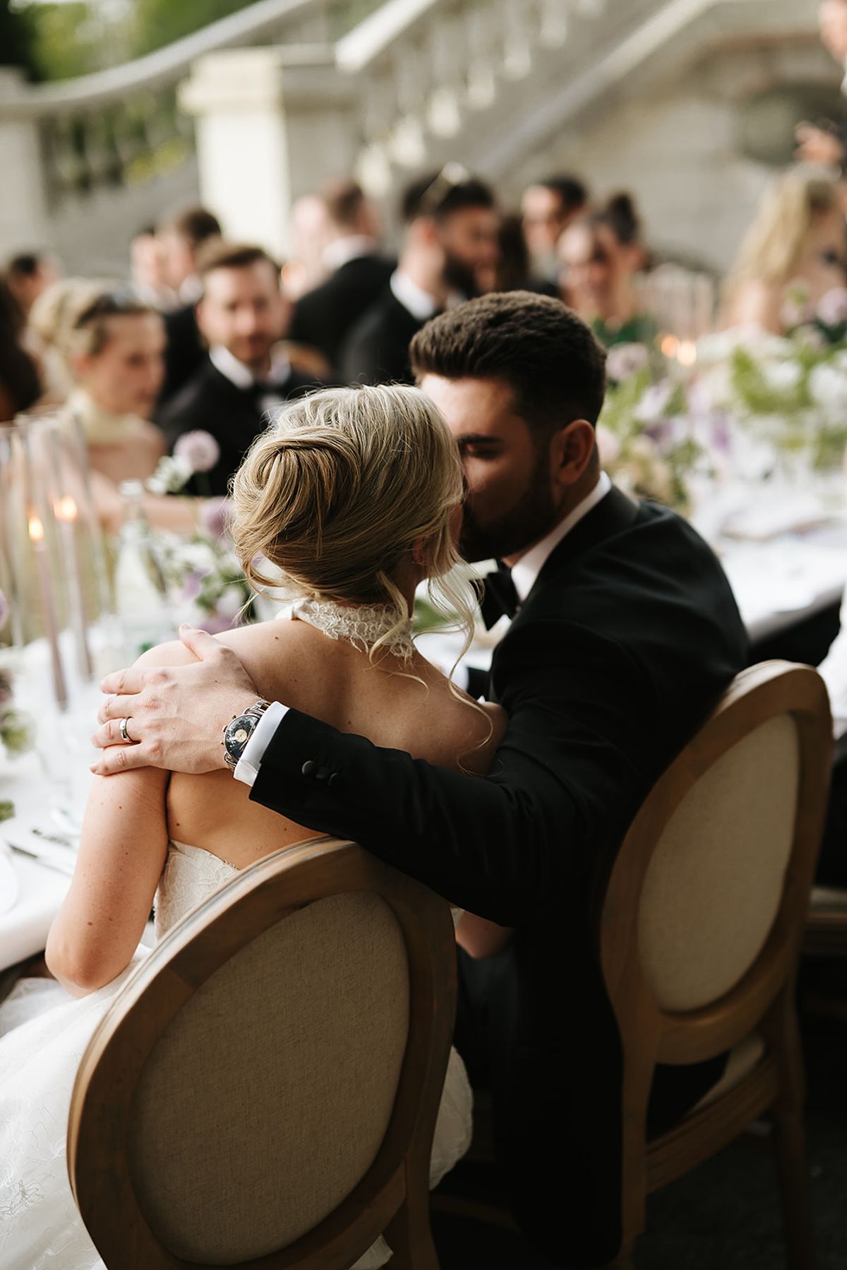 The couple kiss at a long reception table, her in a white lace gown, him in a dark suit, with guests and flowers behind.