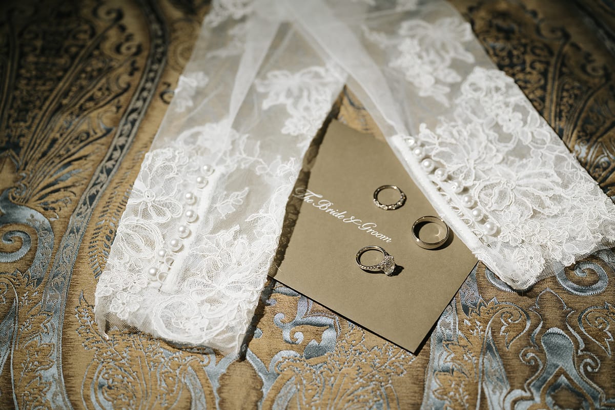 A pair of white lace gloves and two rings on a brown card lie on an ornate patterned fabric.